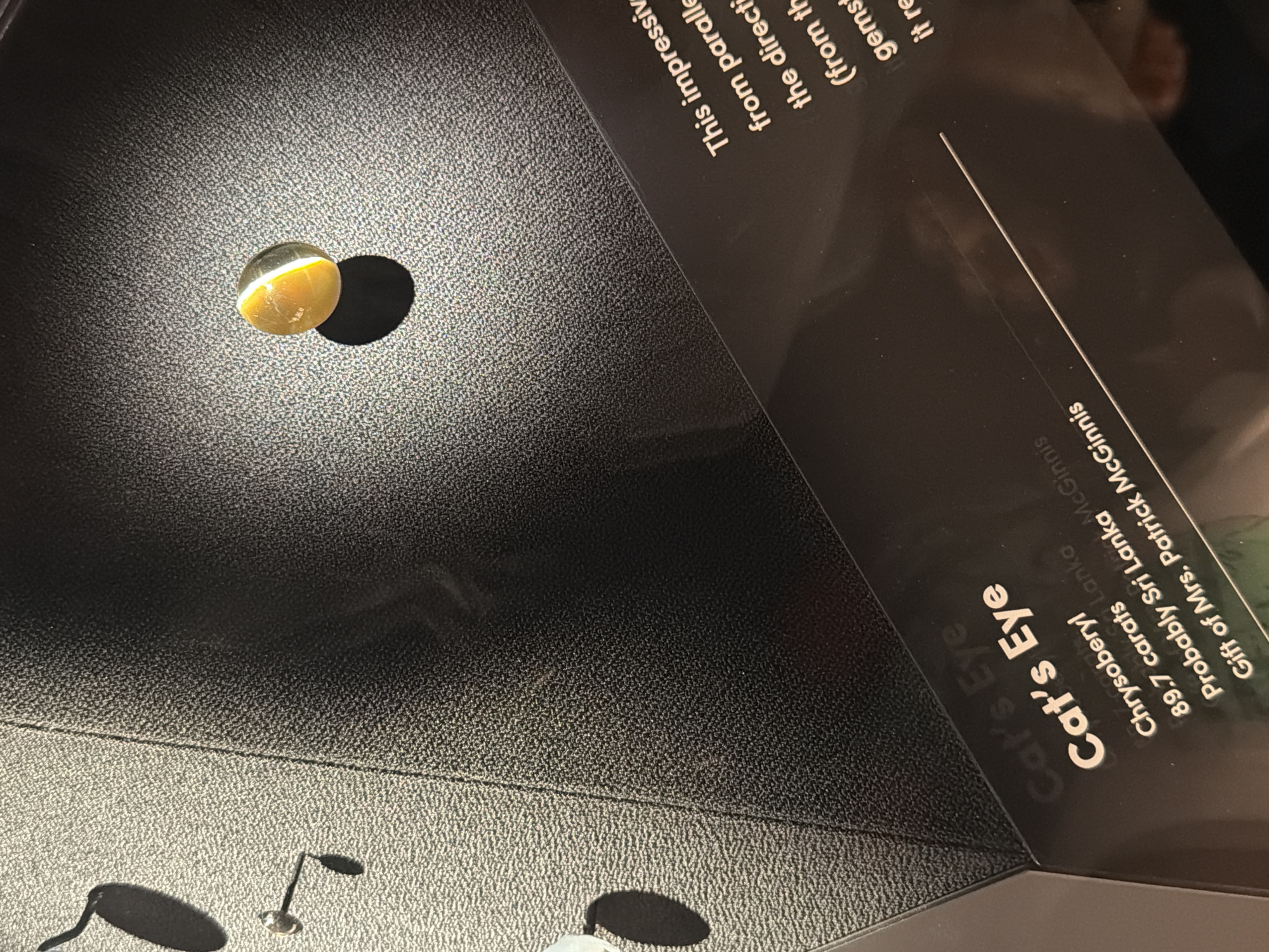 The image shows a museum exhibit featuring a gemstone. The gemstone, labeled "Cat's Eye," is round and appears to be a yellowish-orange color with a distinct white band running through it, giving it a cat's-eye effect. It is displayed on a textured black surface with a dramatic spotlight illuminating it, casting a shadow. A label plate below the gemstone provides further information, stating it is a Chrysoberyl, weighs 89.7 carats, is probably from Sri Lanka, and is a gift of Mrs. Patrick McGinnis. Another label to the right, partially obscured, begins to describe the gemstone's properties. The background consists of angled surfaces with textured black material, and shadows are cast by unseen objects. The scene is devoid of people. The context suggests this is an indoor display in a museum or gallery. There is no information in the image to determine the city or country.