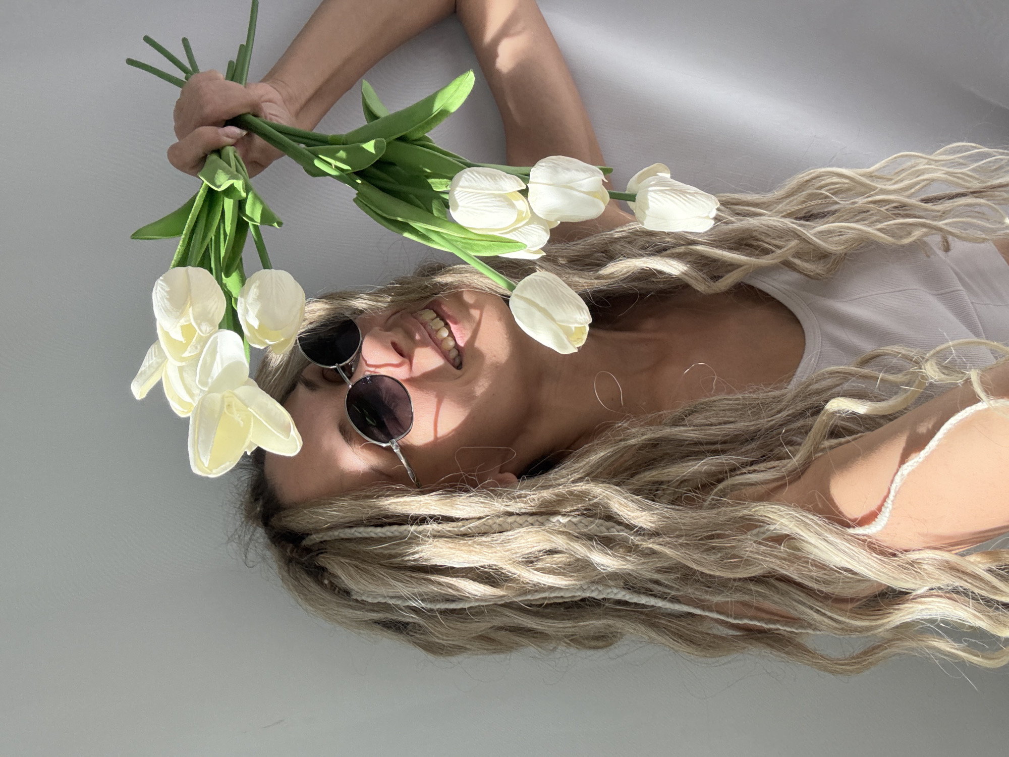 A person is seen from an overhead perspective, lying on their back on a light-colored surface, possibly a floor or bed. The individual has long, wavy blonde hair with several lighter-colored braids or dreadlocks intertwined throughout. They are wearing dark, round sunglasses and a white sleeveless top. Their head is tilted slightly, and a wide, joyful smile is visible beneath the sunglasses, revealing their teeth.
The person's left arm is raised, holding a bouquet of white tulips by their green stems. The tulips have delicate, slightly translucent petals. Strong, directional sunlight casts distinct shadows on the light background, indicating a bright, sunny daytime environment indoors. The overall mood is relaxed and happy. There is no visible text in the image.