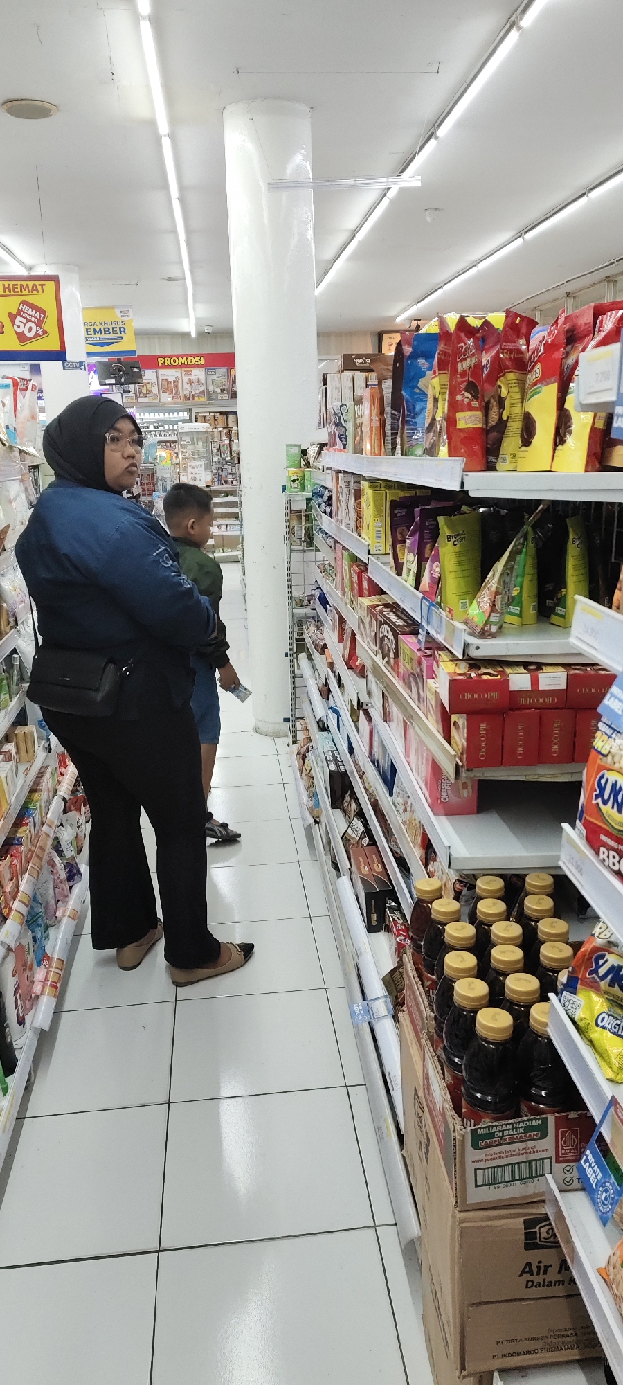 The image shows an aisle in a well-lit convenience store or supermarket. The floor is tiled with large white squares. On the left side of the aisle, a woman wearing a dark blue denim jacket, black hijab, and black pants stands with her left side to the camera, looking to her left. She has a black crossbody bag. A young boy in a green jacket stands to her right and slightly behind her, also looking to the left. He holds a small white object in his right hand.
Shelves are visible on both sides of the aisle, filled with various products, including packaged snacks, drinks, and other goods. Some products are in colorful packaging. On the right side, a prominent display of bottled drinks in a cardboard box is visible, with at least a dozen bottles with yellow caps. Above these drinks, shelves are stocked with boxes and bags of snacks. Signs with Indonesian text are visible on the shelves and walls, indicating promotions and product information. One sign on the left side of the aisle reads "HEMAT 50%".
The overall environment suggests a retail setting, likely within the city of Malang, Indonesia, given the Indonesian language on the signs.