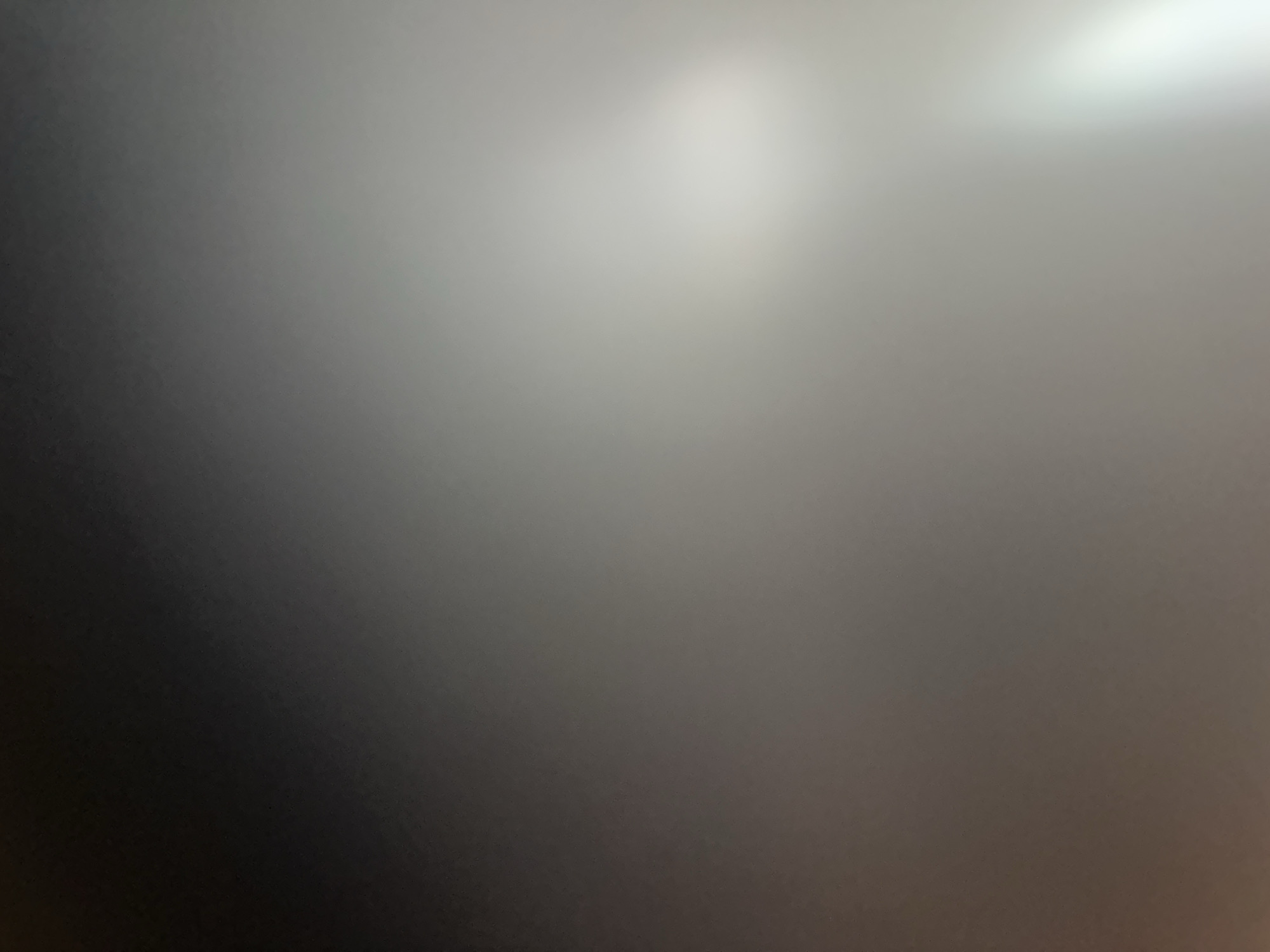 This is a very blurry, abstract image. The dominant colors are shades of gray, transitioning from a dark gray on the left to a bright, washed-out gray or white on the right. There are no discernible subjects like people, objects, or a specific scene. The image appears to be of a surface that is unevenly lit, with a bright light source somewhere off-camera to the upper right, creating a gradient of light and shadow. There is no indication of any activity or event taking place, nor are there any location cues, time of day, weather, or emotional expressions visible. No text is present in the image. Given the lack of distinct features, it's difficult to provide any further context or detail.