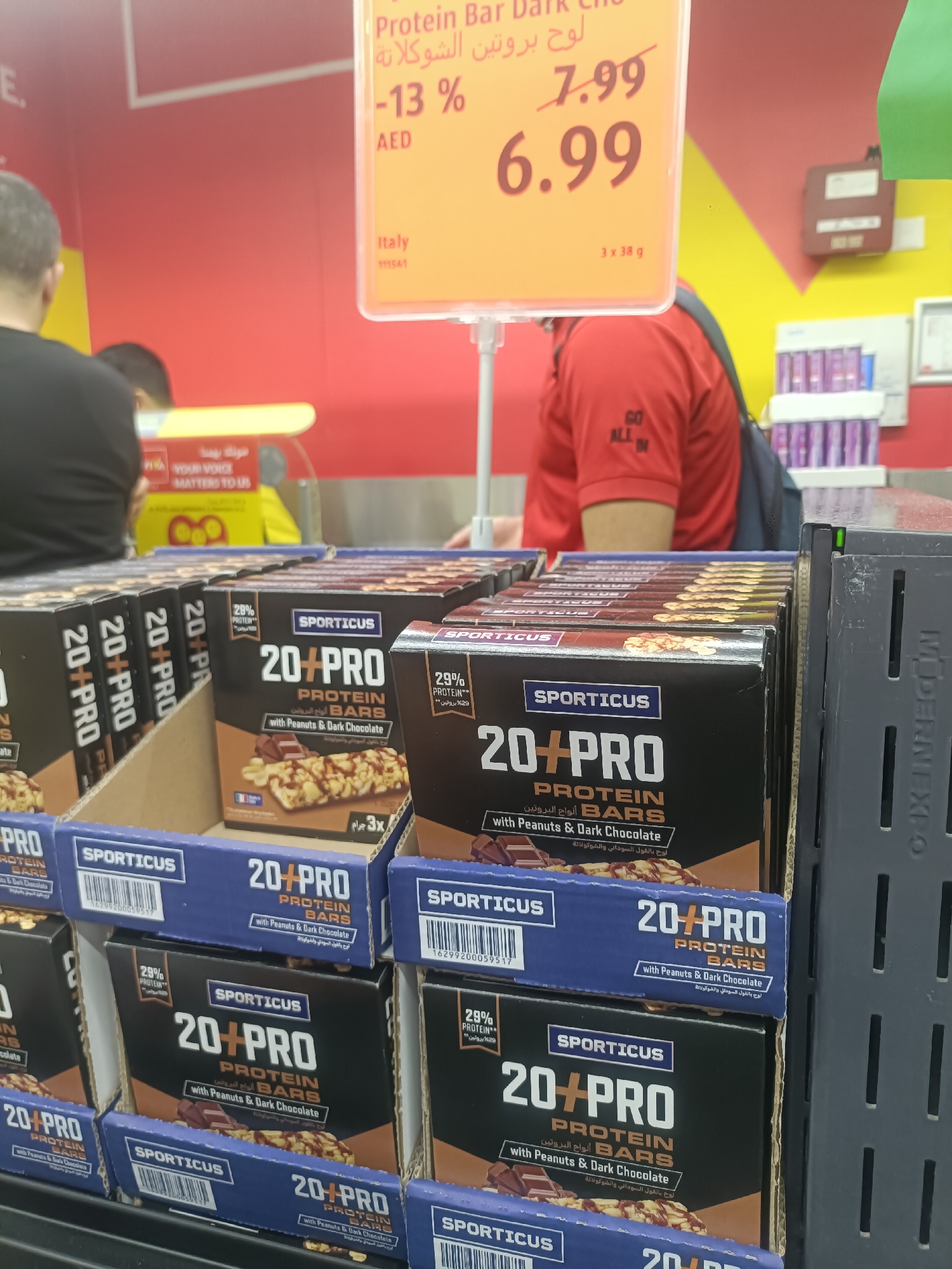 The image shows a retail display of "SPORTICUS 20+PRO PROTEIN BARS with Peanuts & Dark Chocolate" in a supermarket in Dubai, United Arab Emirates. The display features multiple boxes of the product arranged in cardboard trays. A price tag is visible in the upper portion of the image, indicating a sale from 7.99 AED to 6.99 AED. The background shows a red and yellow wall, suggesting a bright, commercial environment. A person wearing a red shirt with the text "GO ALL IN" is partially visible in the background. The products are positioned on shelves, and the overall scene is indicative of a grocery store setting in Dubai.