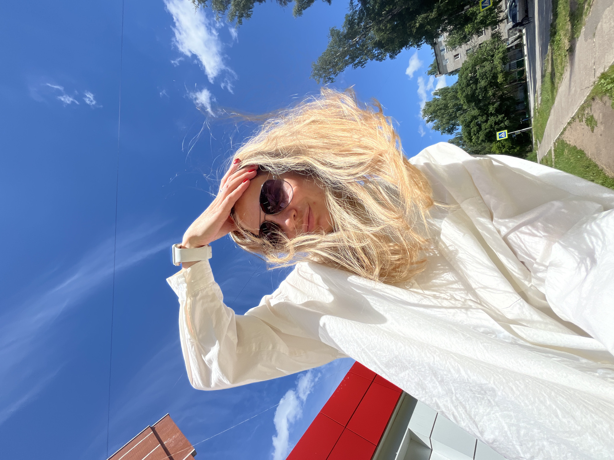 A woman with blonde hair and sunglasses stands outdoors, wearing a white oversized shirt. Her right hand is raised to her head, touching her hair. The background is a bright blue sky with scattered clouds. To the left, a building with red and white sections is visible. To the right, a sidewalk, a street, trees, and a multi-story apartment building are present. A pedestrian crossing sign is visible on the sidewalk. The scene suggests a location in the city of Chaikovsky, Russia.