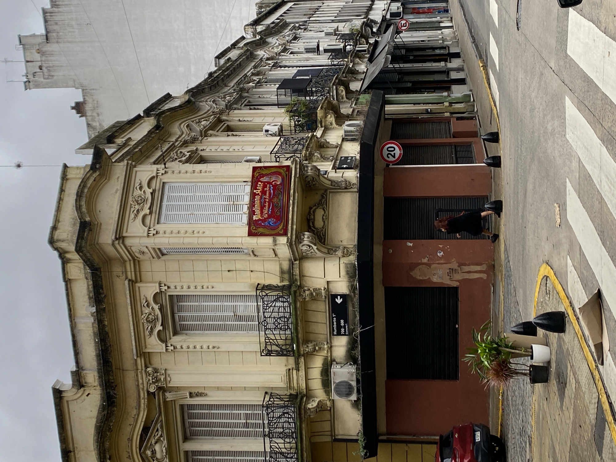 An eye-level, daytime, full shot captures a street scene in Buenos Aires, Argentina. The image is dominated by the ornate façade of a multi-story building with detailed architectural elements, including decorative moldings, wrought iron balconies, and shuttered windows. A sign for "Maelmann Arce" hangs prominently on the building.
To the right of the building, the sidewalk is visible, marked with a zebra crossing. A woman is walking on the sidewalk near a storefront with a closed black shutter. Plants in pots are positioned in front of the store. Street signs are mounted on the side of the building.
The overall impression is one of a historic building in an urban environment, juxtaposed with modern elements like the pedestrian crossing. The sky is overcast, suggesting a cloudy day.