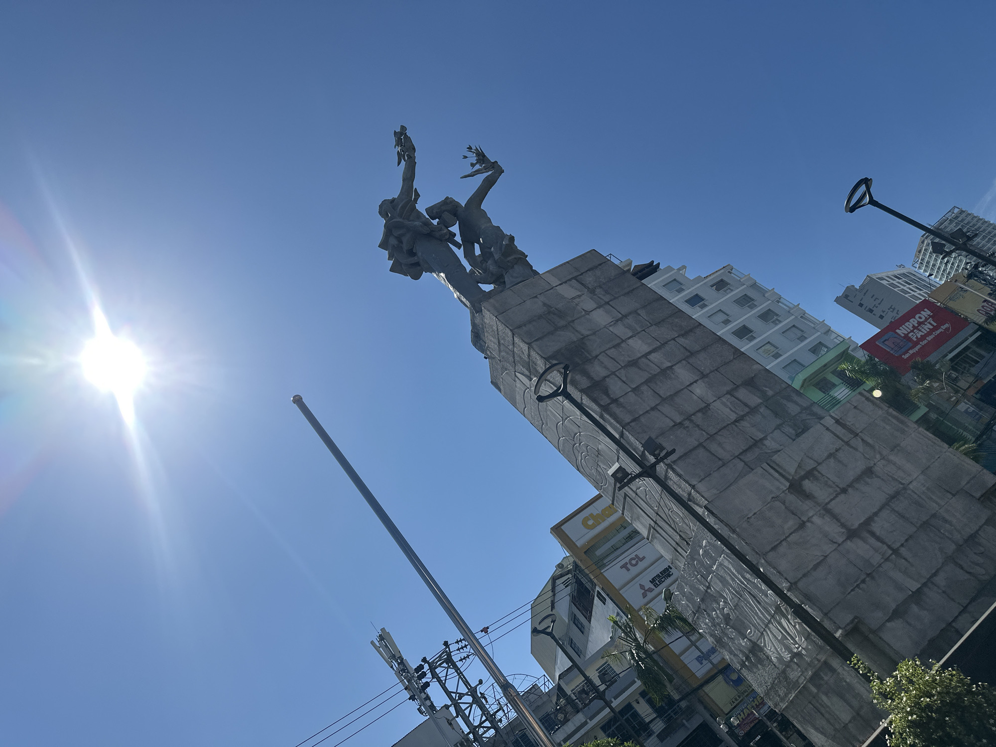 A bright, low-angle, wide shot captures a city scene in Nha Trang Ward, Vietnam, dominated by a large stone monument and a clear blue sky with a strong sun glare. The monument features two bronze figures, one above the other, both reaching forward. The upper figure appears to be male, wearing a backpack, while the lower figure seems to be female, holding a plant. They stand on a grey, segmented stone base.
Behind the monument, a dense collection of buildings rises. Several appear to be commercial establishments, with visible signage for "TCL" and "Mitsubishi Electric." Further back, taller, multi-story buildings create a city skyline. Utility poles with a complex network of wires are prominent on the left side of the image. A tall, slender pole extends upwards from the foreground on the left, its tip obscured by the sun. On the right, a modern lamppost with a circular top stands out against the sky. Lush green foliage is visible at the base of the monument on the left. The overall impression is a bustling urban environment under a very sunny sky.