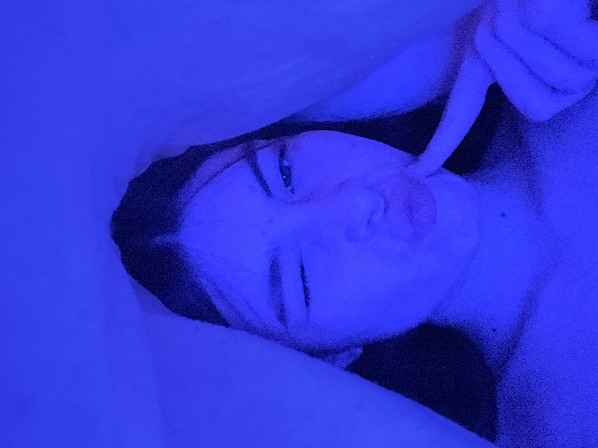 An angled, close-up portrait shows a person, likely female, lying down indoors, illuminated almost entirely by a monochromatic blue light. The light source, possibly an LED lamp or strip, casts a strong, ethereal blue hue across the entire scene, suggesting an evening or nighttime setting.
The person's head is tilted slightly, and they are looking towards the camera. Their left eye appears partially closed, possibly in a playful wink or squint, while their right eye is open. Their lips are gently pursed, contributing to a mischievous or pouting expression. The index finger of their right hand is extended, lightly touching their cheek or jawline. Their dark hair frames their face.
The background is a uniform deep blue, consistent with the pervasive lighting, with vague suggestions of bedding or a pillow supporting the person. The intense blue light creates strong contrasts and highlights, defining the contours of the face while obscuring finer details. There are no distinct objects other than the individual, and no visible text. The overall atmosphere is intimate and stylized by the dramatic lighting. The image captures a moment of playful posing. The location context is Saint Petersburg, Russia.