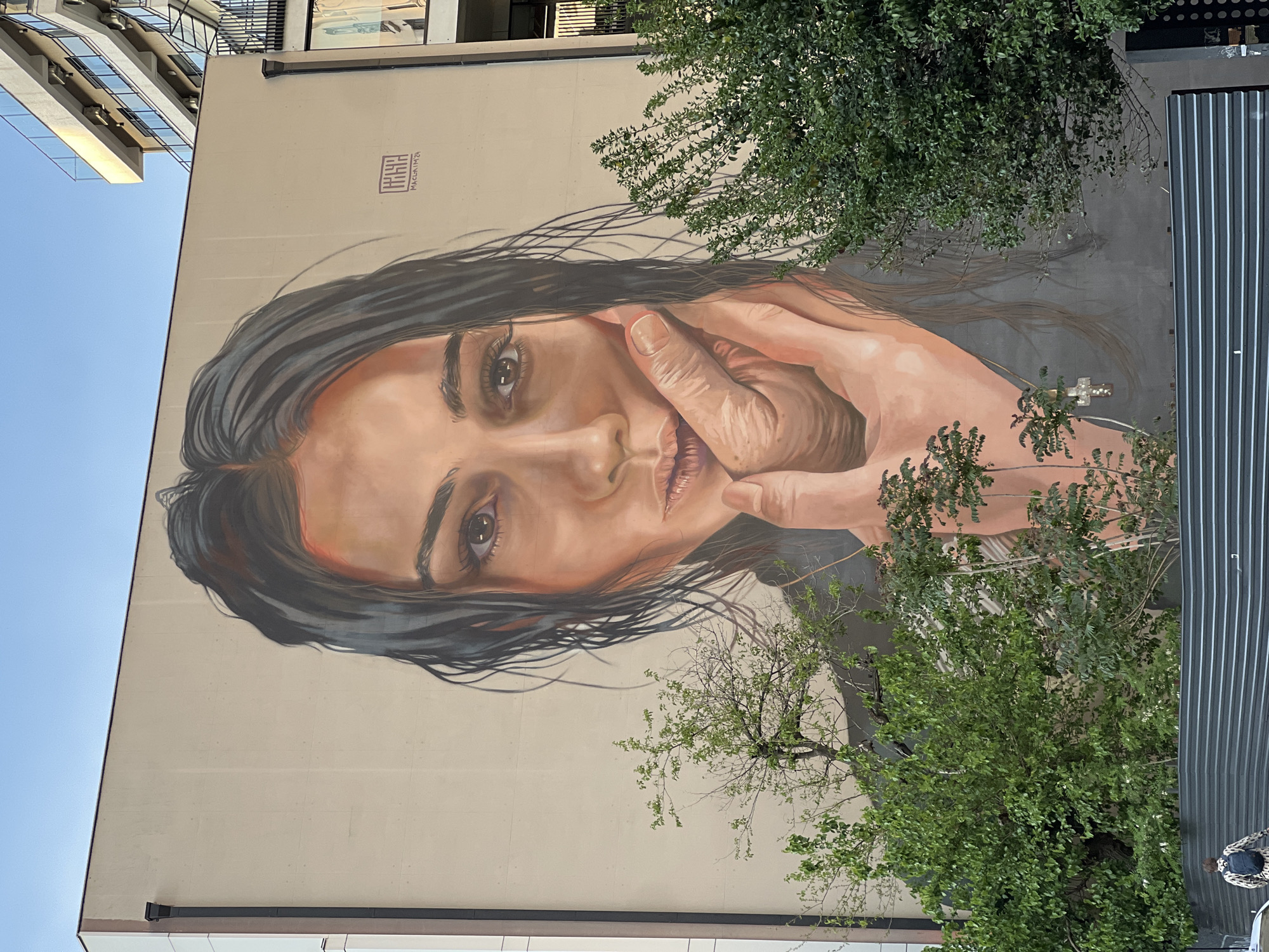 The image shows a large mural on the side of a building. The mural depicts a portrait of a woman with dark hair. She is looking at the viewer with her hand on her face. In the background, there are trees in front of a gray exterior wall. The mural is located in Tbilisi, Georgia.