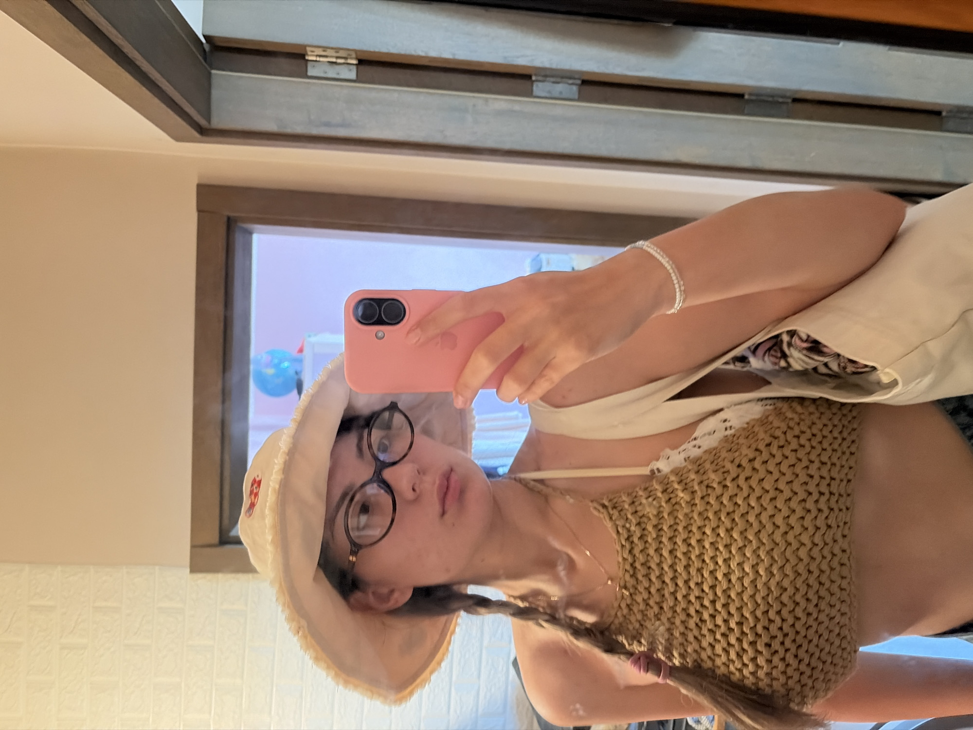 A young woman with braided hair and glasses is taking a mirror selfie in what appears to be a room with a brick-patterned wall and a wooden door. She is wearing a wide-brimmed hat, a knitted top, and a white bra. She is holding a pink smartphone in her right hand and a white tote bag is slung over her left shoulder. The setting does not provide any identifiable visual cues to suggest a specific location within Tsarevo, Bulgaria.