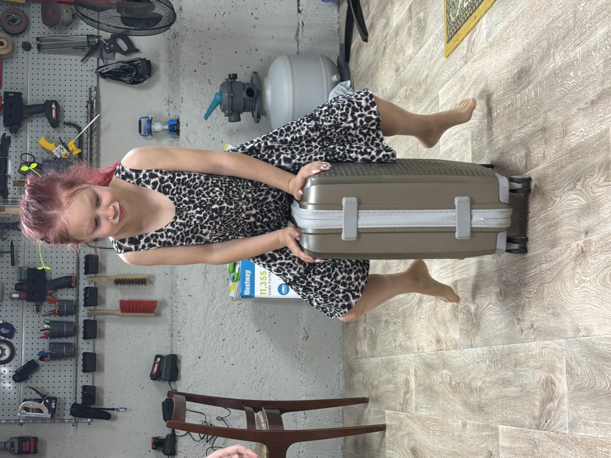 A young female with dyed red hair sits on a silver suitcase indoors, possibly in a garage or basement. The wall behind her features a pegboard with various tools and hardware attached. A water tank is mounted above the pegboard, and a Bestway sand filter box sits beside the suitcase. The floor is light-colored wood-look laminate, and a partially visible wooden chair is present in the foreground. The location in Dubovaya, Russia, is not evident from the visual content.