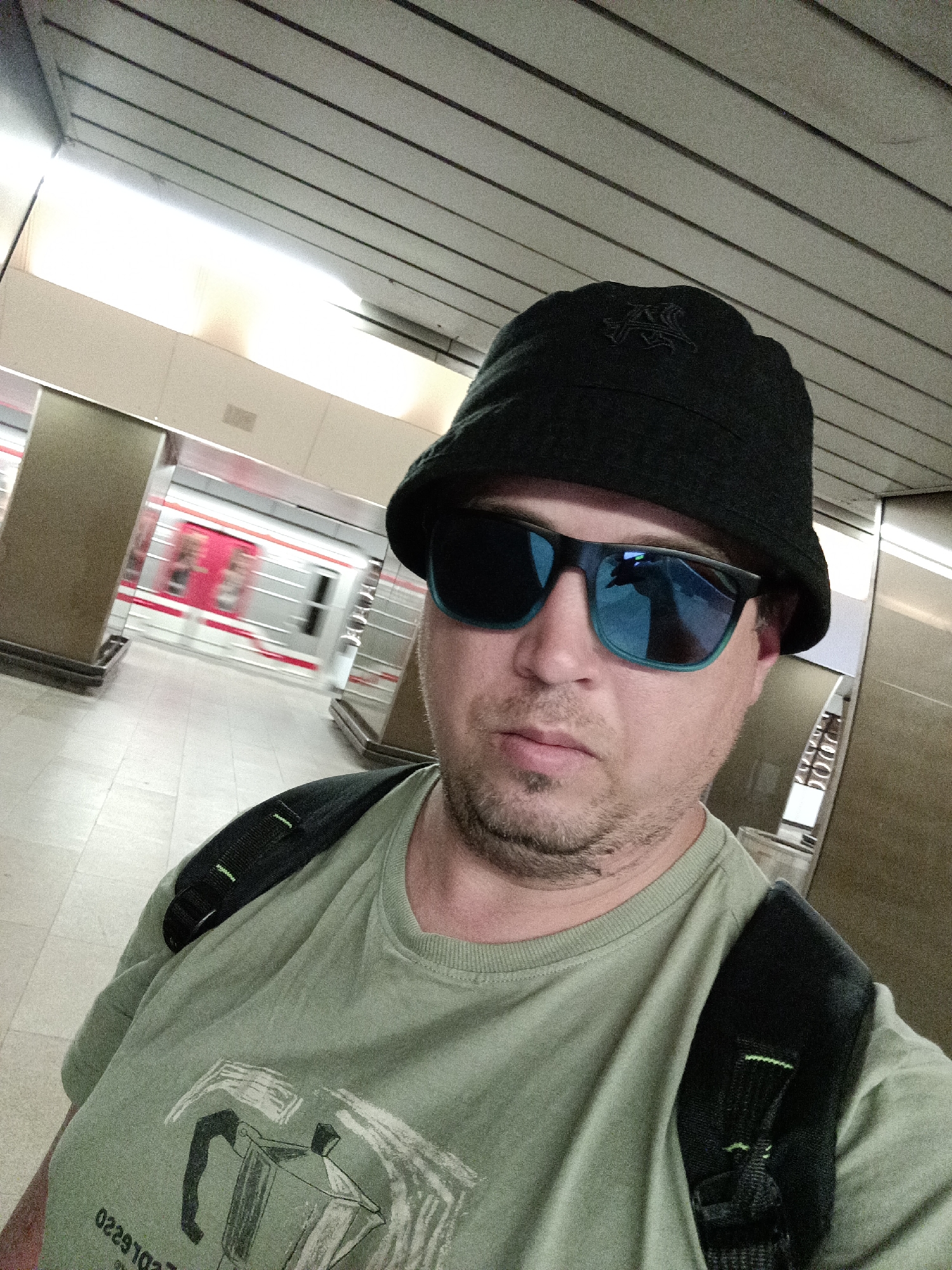 A man wearing a black bucket hat, sunglasses, and a green t-shirt with a graphic print is standing on a subway platform. He is looking at the camera, with a train blurred in motion behind him. The platform has tiled flooring and modern architectural elements with lighting fixtures on the ceiling. This image was taken in Prague, Czechia.