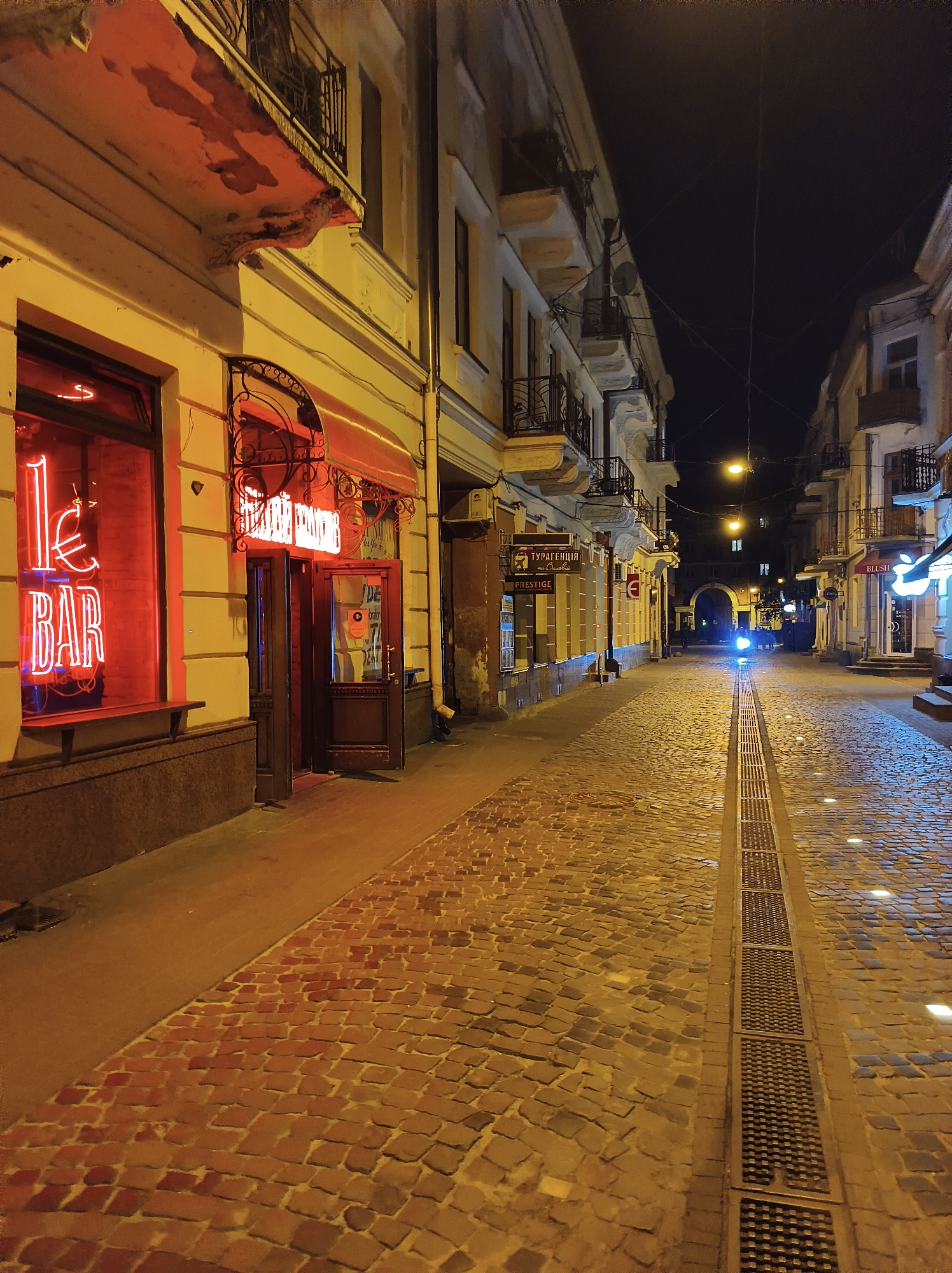 The image shows a narrow cobblestone street in Ternopil, Ukraine at night. The street is lined with buildings on both sides, featuring balconies and ornate architectural details. A neon sign that reads "LE BAR" in red letters is prominently displayed on the left side of the street, illuminating the facade of a building. An open doorway with a red awning above it suggests an establishment is active. The cobblestone pavement is illuminated by streetlights and the glow from shop windows. A drainage grate runs down the center of the street. In the distance, an arched passageway is visible, with a single bright light shining through it. The scene is deserted, conveying a quiet, nocturnal atmosphere.