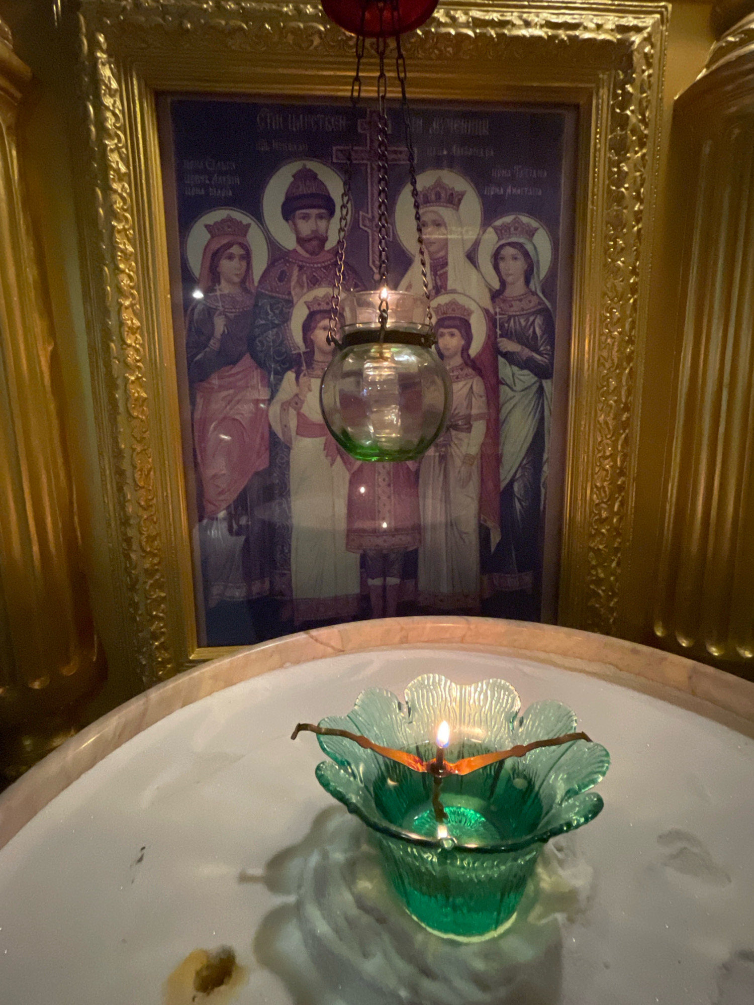 In Pattaya City, Thailand, an Orthodox icon depicting a family, likely the Romanovs, is displayed within a gold ornate frame. The icon shows a man and woman, presumably parents, with three children. To the left of the icon, a golden fluted column is visible. In the foreground, a green glass oil lamp, shaped like a flower and filled with oil, burns with a single flame. The lamp is suspended by chains and is placed within a white basin, possibly for holy water.