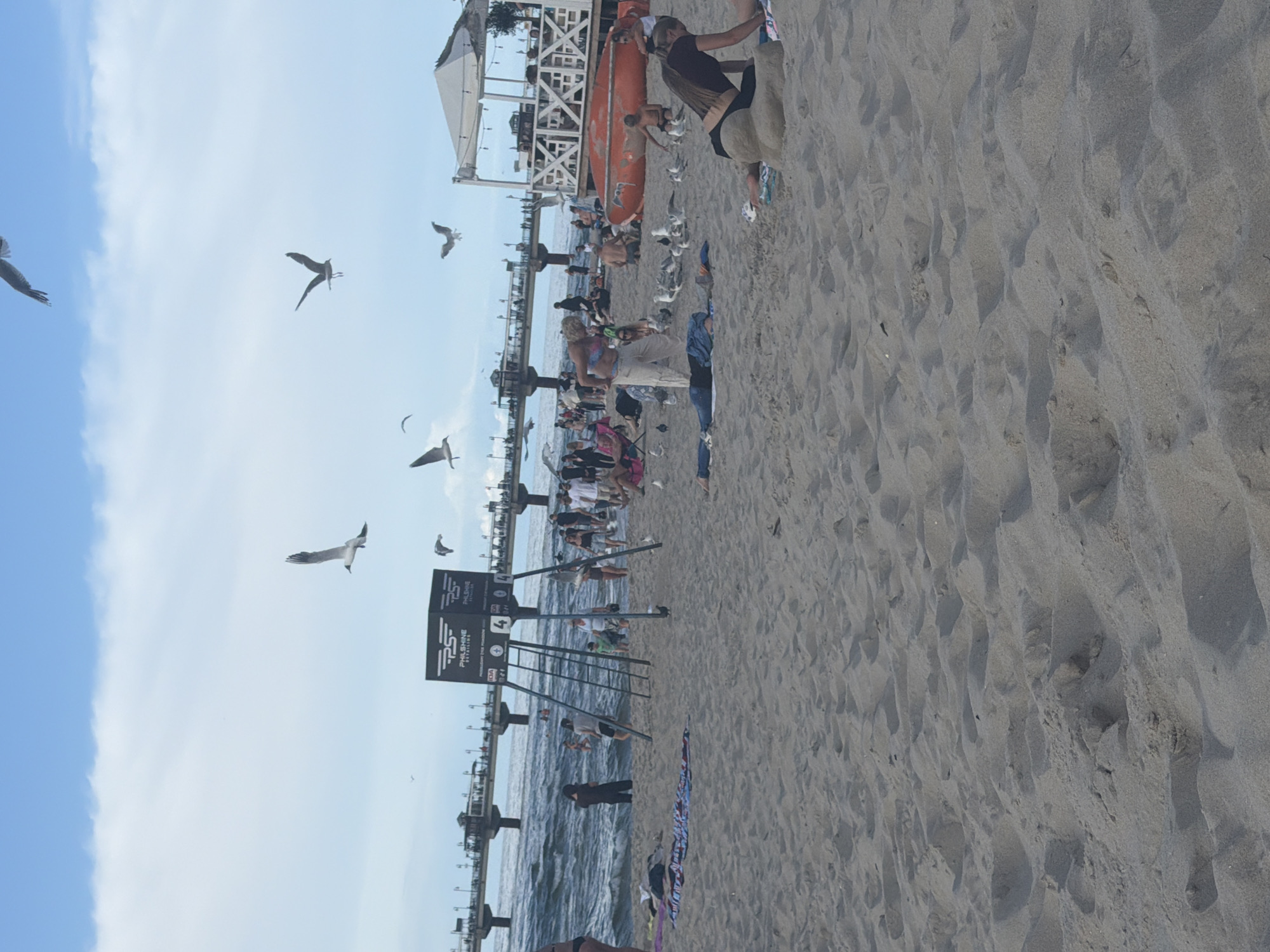 The image depicts a lively scene at the beach in Międzyzdroje, Poland. The foreground is dominated by sandy terrain with footprints and shadows from the midday sun. A woman in a brown top and black bikini bottoms is positioned on the right, appearing to be sitting on the sand. In the mid-ground, the beach is populated with numerous people engaging in various activities. Some are walking, others are sitting or lying on towels, and a group of individuals are interacting near the water's edge. A pier extends into the sea in the background, with a white, open-air structure that resembles a beach bar or restaurant located to the right of the pier. Several seagulls are visible in the sky, with one in flight near the top left of the frame. The sky is partly cloudy, with a mix of blue sky and white clouds. A black and white sign with "PHILSHINE DETAILING" branding is mounted on a tall, metal structure near the center of the mid-ground. An orange, boat-like object is situated on the sand near the white structure. The ocean waves are visible, and the overall atmosphere appears to be one of a busy summer day at the coast.