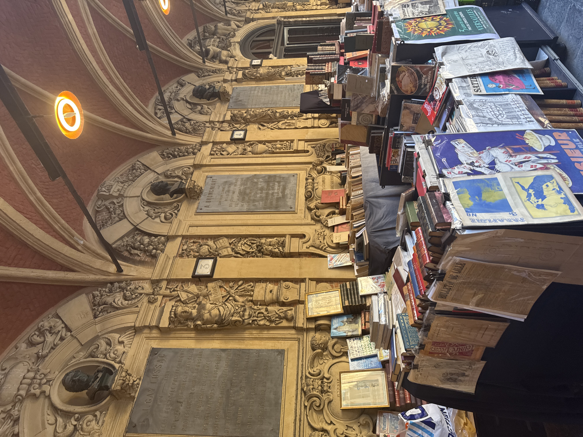 This image depicts an outdoor book stall set up under a covered arcade with arched ceilings and ornate architectural detailing. The stall displays a large quantity of books, framed prints, and papers. The books are arranged on tables and shelves, with some stacked high and others neatly lined up. Various types of paper items, including what appear to be newspapers and posters, are hung from clips.
The architectural style suggests a historical European setting. The inscription on a plaque visible in the upper left reads "J. GAY LUSSAC", indicating a possible connection to scientific or academic history. Another plaque mentions "A. BRONNIART". These inscriptions, along with the overall aesthetic of the arcade, strongly suggest a location within the city of Lille, France, potentially in a historically significant area such as the Grand' Place or near the Palais des Beaux-Arts, which are known for their arcades and historical architecture.
There are no visible people in the immediate foreground, but the presence of the book stall implies an active marketplace or a vendor preparing for business. The lighting suggests it is daytime, with natural light filtering in from the open ends of the arcade and artificial lights visible on the ceiling. The environment is orderly, with the merchandise carefully displayed. The focus is on the books and antique-like items, evoking a sense of history and culture.