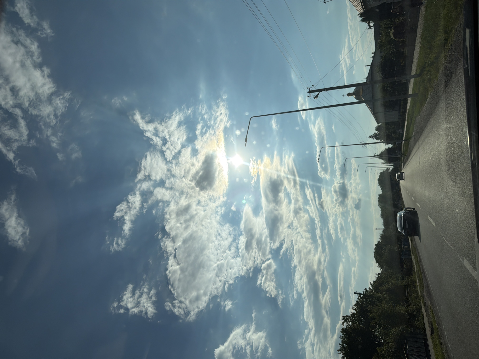 The image shows a road with trees lining one side and buildings on the other, under a partly cloudy blue sky with the sun shining brightly. A black car is driving on the road in the foreground, and another car is visible in the distance. There are power lines and streetlights along the road. The architecture of the buildings suggests a location in a village or small town. Specifically, a domed building resembling a church is visible in the background. The overall scene depicts a sunny day on a rural road in Teofipilka, Ukraine.