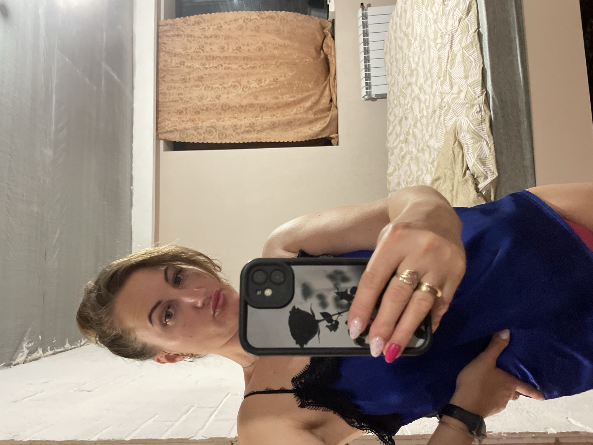 A woman is taking a mirror selfie in a dimly lit room. She has blonde hair pulled up in a bun and is wearing a blue slip with black lace trim. She is wearing rings on her fingers and a watch on her left wrist. In the background, there is a bed with patterned bedding, a window with a curtain, and a radiator. The ceiling appears to be unfinished concrete. The location is not specified as Zarichne, Ukraine.