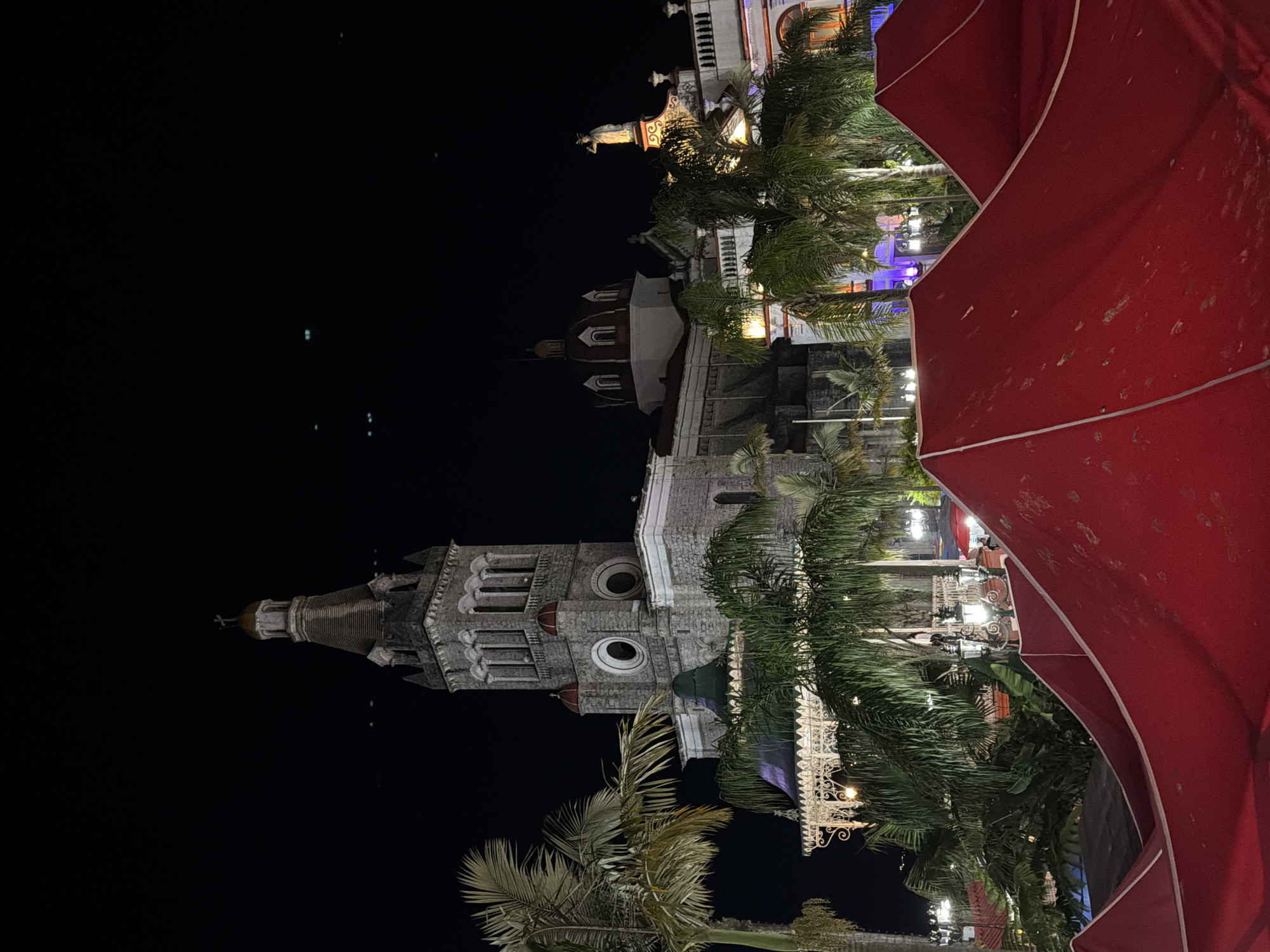 The image depicts a night view of the Iglesia de San Francisco in Cuetzalan, Mexico. The church's facade, with its distinctive tower and ornate details, is prominently featured against a dark sky. Palm trees are interspersed around the building, suggesting a tropical environment. Part of a red umbrella is visible in the foreground, adding a foreground element to the shot. The image suggests a nighttime urban scene with a focus on a significant architectural landmark.