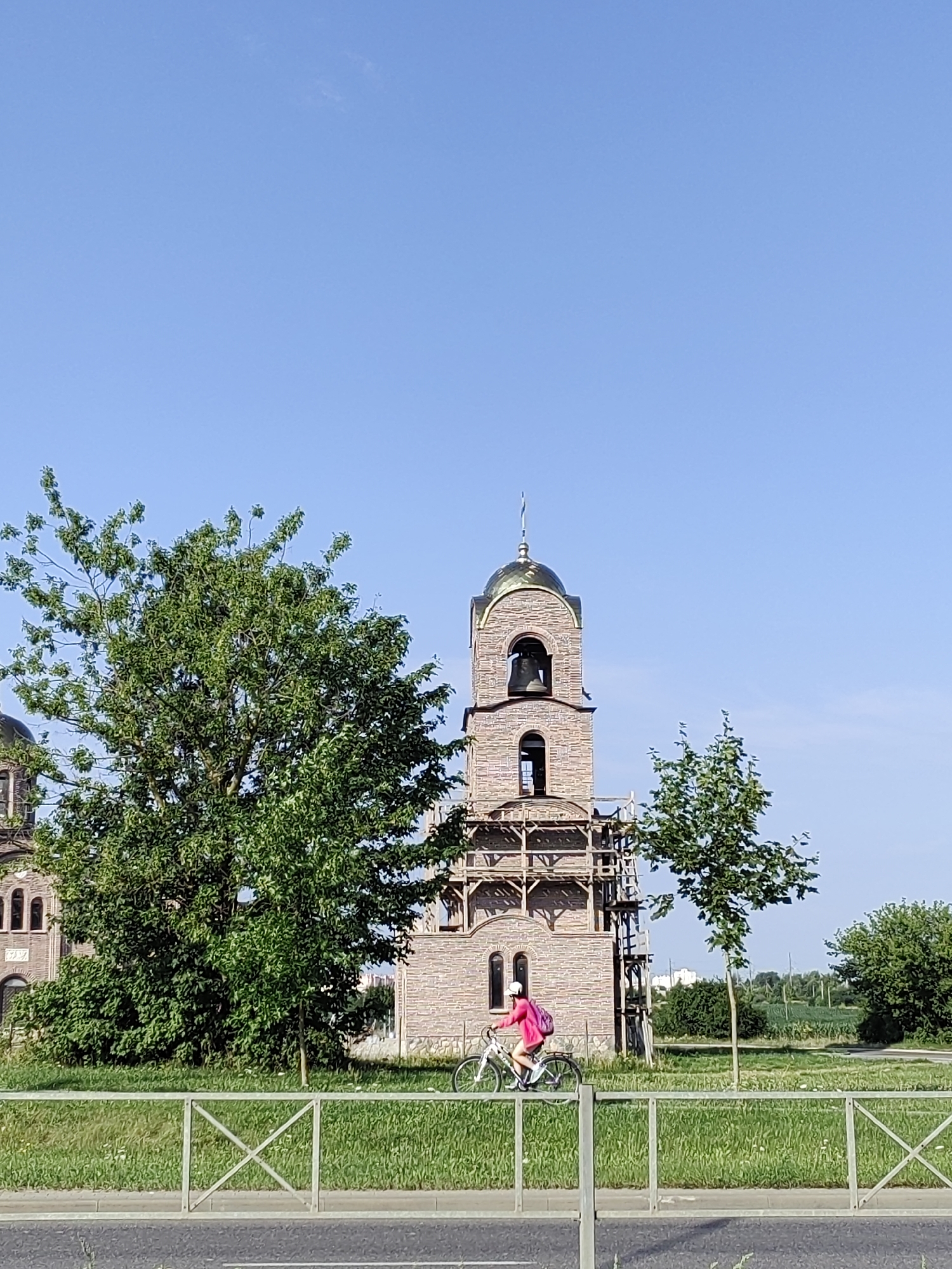 The image depicts a scene in Hrodna, Belarus, featuring an Orthodox church under construction. A young girl with a pink jacket and backpack is riding a bicycle on a paved path next to a grassy area. The church's bell tower, adorned with a golden dome and a cross, is partially visible. The construction site is surrounded by scaffolding. The background shows lush green trees and fields, with a clear blue sky above. A metal fence runs along the foreground, separating the path from the road.