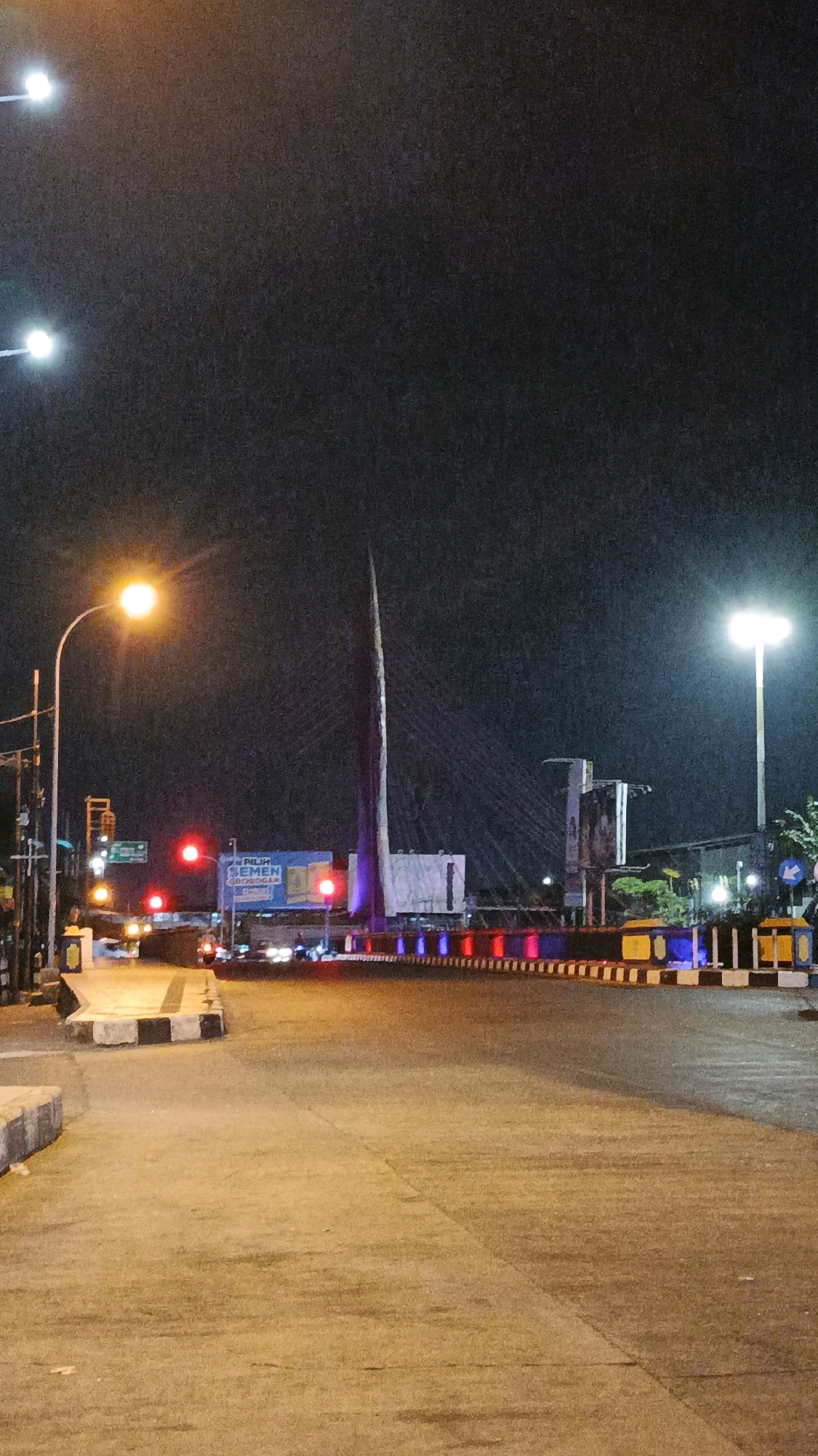 The image depicts a nighttime view of a street in Surakarta, Indonesia, likely near the Jl. Slamet Riyadi overpass. The foreground shows a paved road with street lighting reflecting on its surface. In the midground, a traffic light displays a red signal. Further back, a distinctive cable-stayed bridge is visible, its tall pylon illuminated. The bridge is adorned with a series of colorful lights, alternating between red, blue, and yellow, which run along its railing. On the right side of the frame, a lamppost casts a bright light on the scene. Several vehicles are present on the road, their headlights and taillights visible. A billboard with text in Indonesian, "PILIH SEMEN GRESIC", is situated behind the traffic light. The sky is dark, indicative of nighttime. The overall scene captures a typical urban environment at night with illumination from streetlights, vehicles, and decorative bridge lighting.