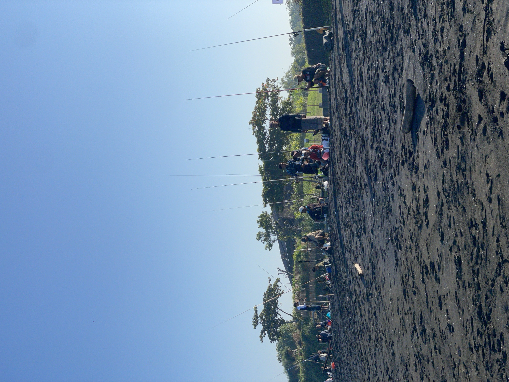 The image depicts a group of people fishing on a black sand beach with a clear blue sky overhead. The individuals are spaced out along the shoreline, each with a fishing rod. Some are sitting, while others are standing or crouching. In the background, there is a line of trees and vegetation, and a small building or structure is visible. The overall impression is a calm, outdoor activity taking place on a sunny day. The location is specified as Kediri, Indonesia.