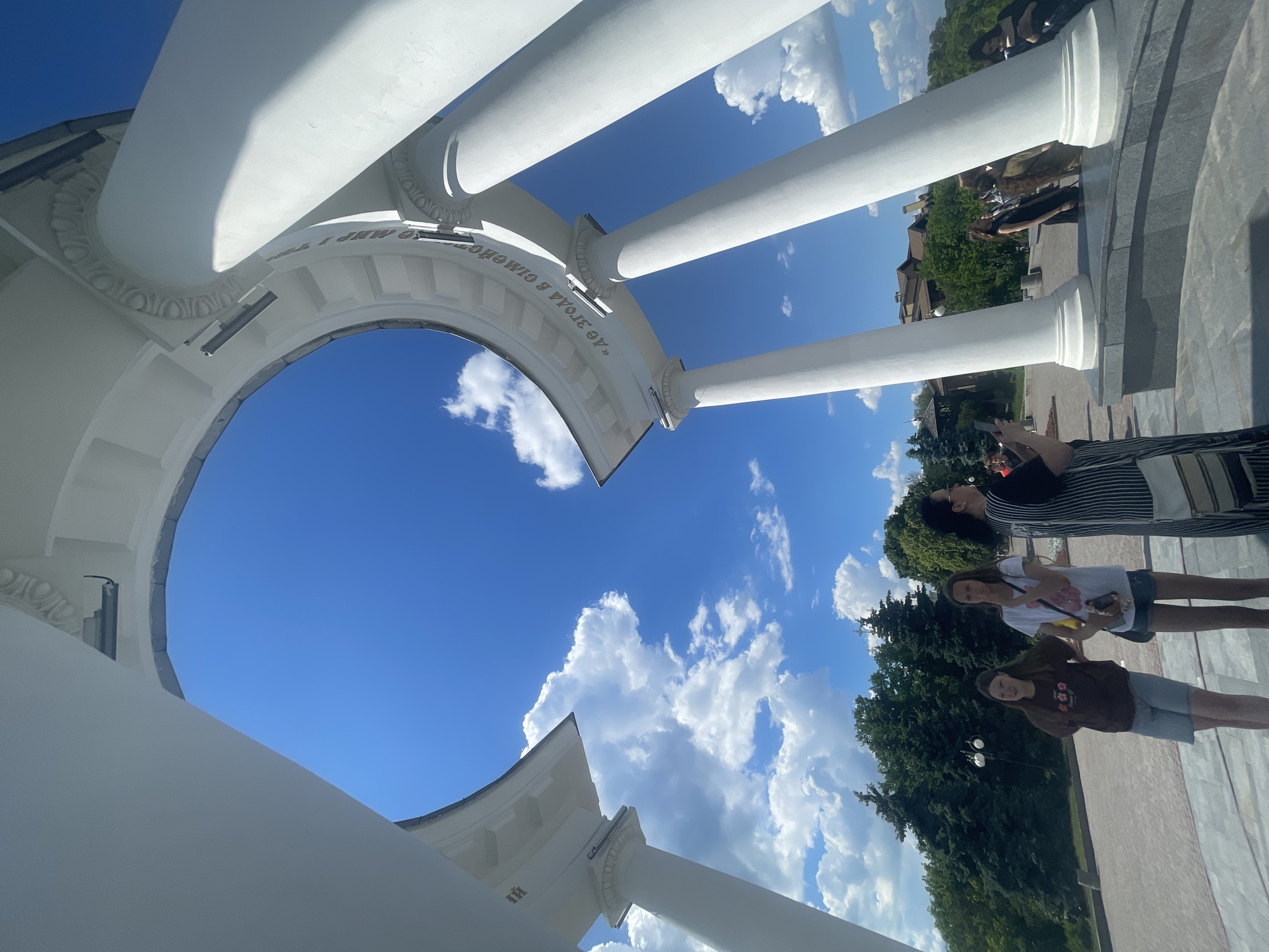 The image, correctly oriented, presents an upward view through a bright white, classical-style architectural structure, likely a rotunda or colonnade, set against a vibrant blue sky filled with scattered white cumulus clouds. The structure is composed of sturdy white columns supporting arches, adorned with decorative elements resembling egg-and-dart moulding along the frieze.
Three main individuals are visible, standing on paved ground beneath the architectural feature. In the foreground, a woman wearing a black and white striped top holds a smartphone, seemingly capturing a photo or video of the surroundings. To her right, a girl in a white t-shirt and blue shorts holds a small, light-colored object. Further right, another girl in a brown long-sleeved top and denim shorts stands facing the view. Lush green trees are visible in the background, suggesting a park or public garden setting.
The scene takes place during bright daylight, indicative of sunny weather. The people appear to be casually visiting the site, enjoying the architecture and the view. A notable detail is the partial inscription in Cyrillic script visible on the inner surface of one of the arches: "«ДЕ ЗГОДА Й СИМВОЛ" (meaning "Where there is agreement and a symbol"). This phrase is a strong identifier for the White Arbor (Біла альтанка) or Rotunda of Friendship, a well-known landmark in Poltava, Ukraine.