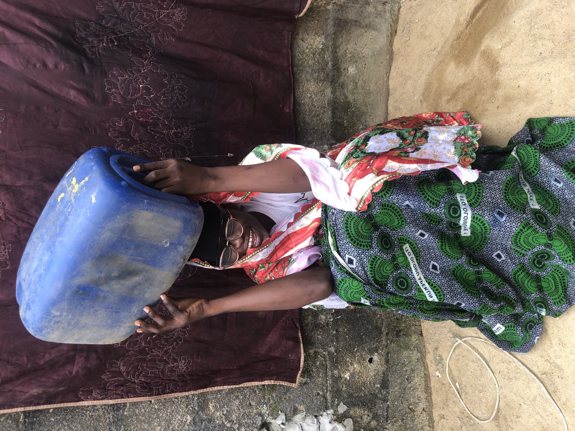 A dark-skinned person wearing a black headscarf, glasses, and a colorful patterned outfit is sitting on the ground, balancing a large blue plastic container on their head. The person is smiling. They are seated in front of a wall that appears to be made of rough concrete or stone, with a dark maroon fabric hanging behind them, embroidered with a white floral pattern. The ground is sandy or dirt. A white rope is coiled on the ground to the left of the person. The scene appears to be outdoors in Ado, Nigeria, possibly in a residential or communal area where water is being transported.