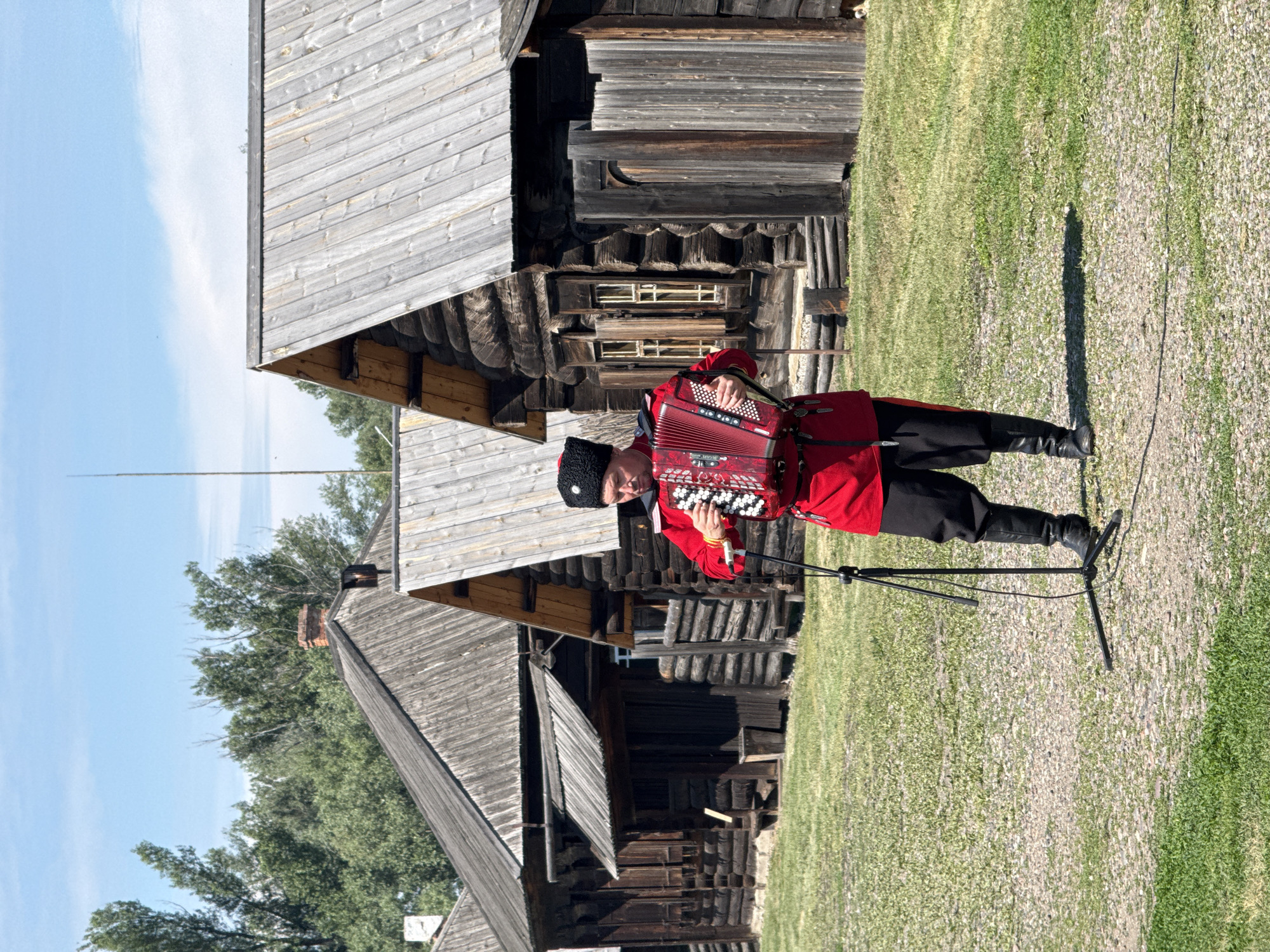 In Shushenskoye, Russia, a man in traditional Cossack attire, including a red tunic, black trousers, and a black fur hat, plays a red accordion. He stands in a grassy area in front of several wooden log houses. A microphone on a stand is positioned in front of him. The sky is blue with some clouds, and trees are visible in the background. The log houses have wooden roofs and appear to be old structures.