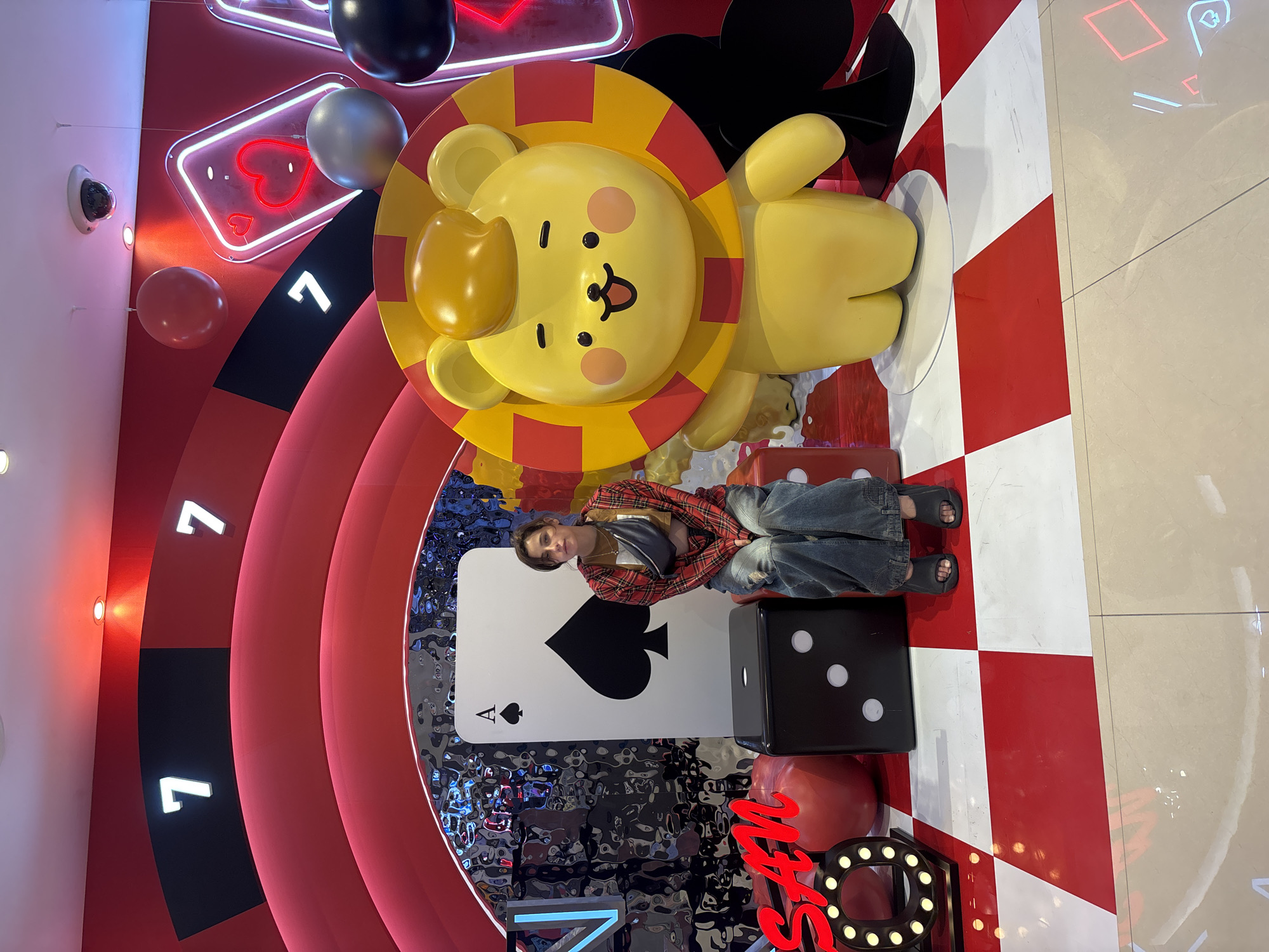 The image captures a promotional display likely in Busan, South Korea, featuring a casino-themed setup. In the center, a young person, dressed in a plaid shirt, a cropped tan top, and baggy jeans, is seated on a large black die. Their arms are resting on their knees, and their gaze is directed slightly to the left.
The backdrop is dominated by a large, stylized roulette wheel structure with illuminated "7"s. Neon signs shaped like playing cards, including a heart and a diamond, add to the vibrant, playful atmosphere. A large, cheerful yellow cartoon lion statue stands to the right of the person. The floor is a striking red and white checkered pattern. Scattered around the display are other casino-related elements, such as large red balls and a giant ace of spades card. The overall environment suggests an indoor commercial space, possibly a mall or a themed event area.