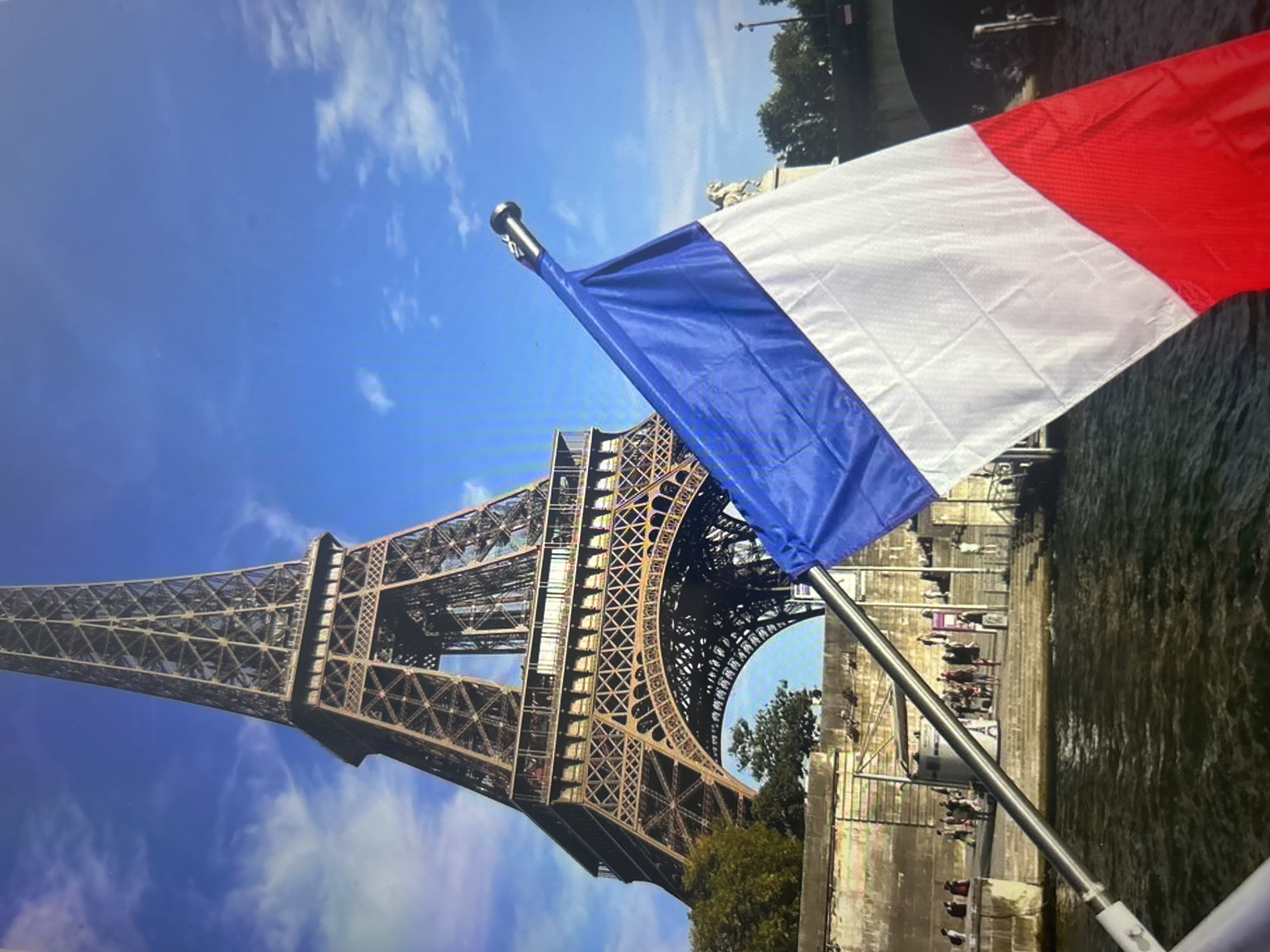 The image captures a vibrant daytime scene in Paris, France, showcasing a prominent French flag and the iconic Eiffel Tower. The French flag, with its vertical blue, white, and red stripes, is positioned in the foreground, attached to a silvery pole that extends from the lower right of the frame. Its placement suggests the photo was taken from a boat or a viewing platform near the water.
In the mid-ground, the magnificent iron structure of the Eiffel Tower dominates the left side of the composition, reaching high into a clear blue sky dotted with soft white clouds. Its intricate lattice design is clearly visible. Below the flag and extending across the bottom right, a body of water, likely the River Seine, flows calmly. Along the riverbanks, stone walls and pathways are discernible, populated by numerous small figures of people walking or gathering, indicative of a bustling tourist area. Green trees and other urban structures line the river's edge.
The scene depicts a typical sightseeing experience in the French capital. The bright blue sky and the presence of scattered clouds indicate fair and sunny weather, perfect for outdoor activities. The Eiffel Tower and the French flag serve as strong location cues, immediately identifying Paris. No readable text is visible in the image.