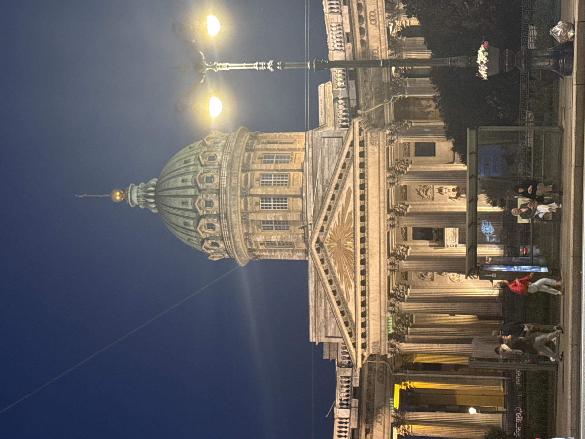 The image captures the Kazan Cathedral in Saint Petersburg, Russia, at dusk. The grand neoclassical cathedral, with its iconic colonnade and dome, is illuminated by artificial lights, casting a warm glow on its facade. A streetlamp with two glowing bulbs stands prominently in the foreground on the right, its ornate design adding to the historical ambiance. In front of the cathedral, a bus stop shelter is visible, with a few people waiting. Some individuals are walking along the sidewalk, while others are seated inside the shelter. The night sky is a deep, clear blue, with faint wisps of clouds or contrails stretching across it. The scene depicts a blend of historical architecture and modern urban life, with the cathedral serving as a majestic backdrop to the everyday activities of the city.