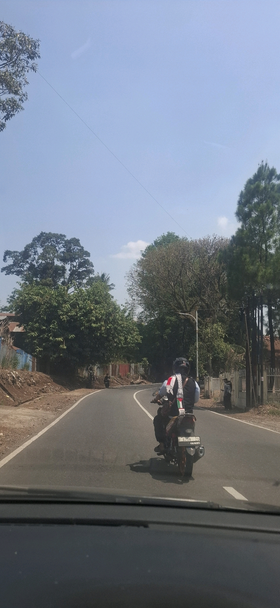 The image depicts a sunlit, paved road curving to the left, with a clear blue sky above and abundant green foliage bordering the road. A person is riding a motorcycle away from the viewer on the right side of the road. The motorcycle has a license plate with the characters "F 6208 ZD". The rider is wearing a helmet and a vest over their clothing. In the background, there are trees, a light pole, and some buildings with corrugated metal siding. The road appears to be in a semi-rural or suburban area. The presence of the license plate format "F" suggests the location is in West Java, Indonesia, and the context of Cilaku, Indonesia, is given.