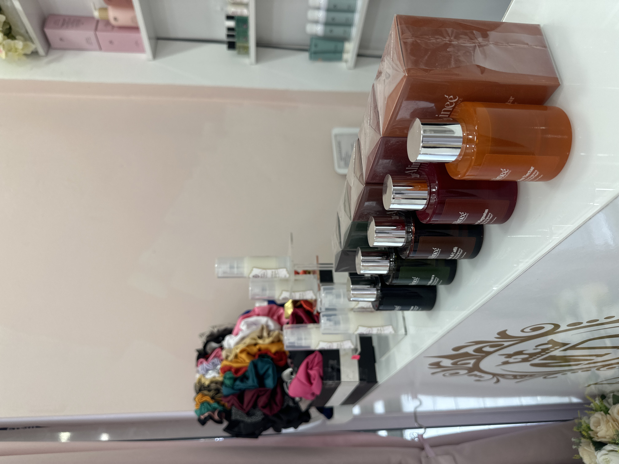 The image shows a shelf stocked with various hair care products and scrunchies. The products, a brand called "Alinee", include boxed sets and individual bottles in shades of brown, red, green, and black. The bottles are neatly arranged in a row. Adjacent to the hair care products, a collection of colorful scrunchies is displayed on a tiered stand. The shelf itself is white with a decorative gold-colored pattern. The location of this image is not discernible from the given context.