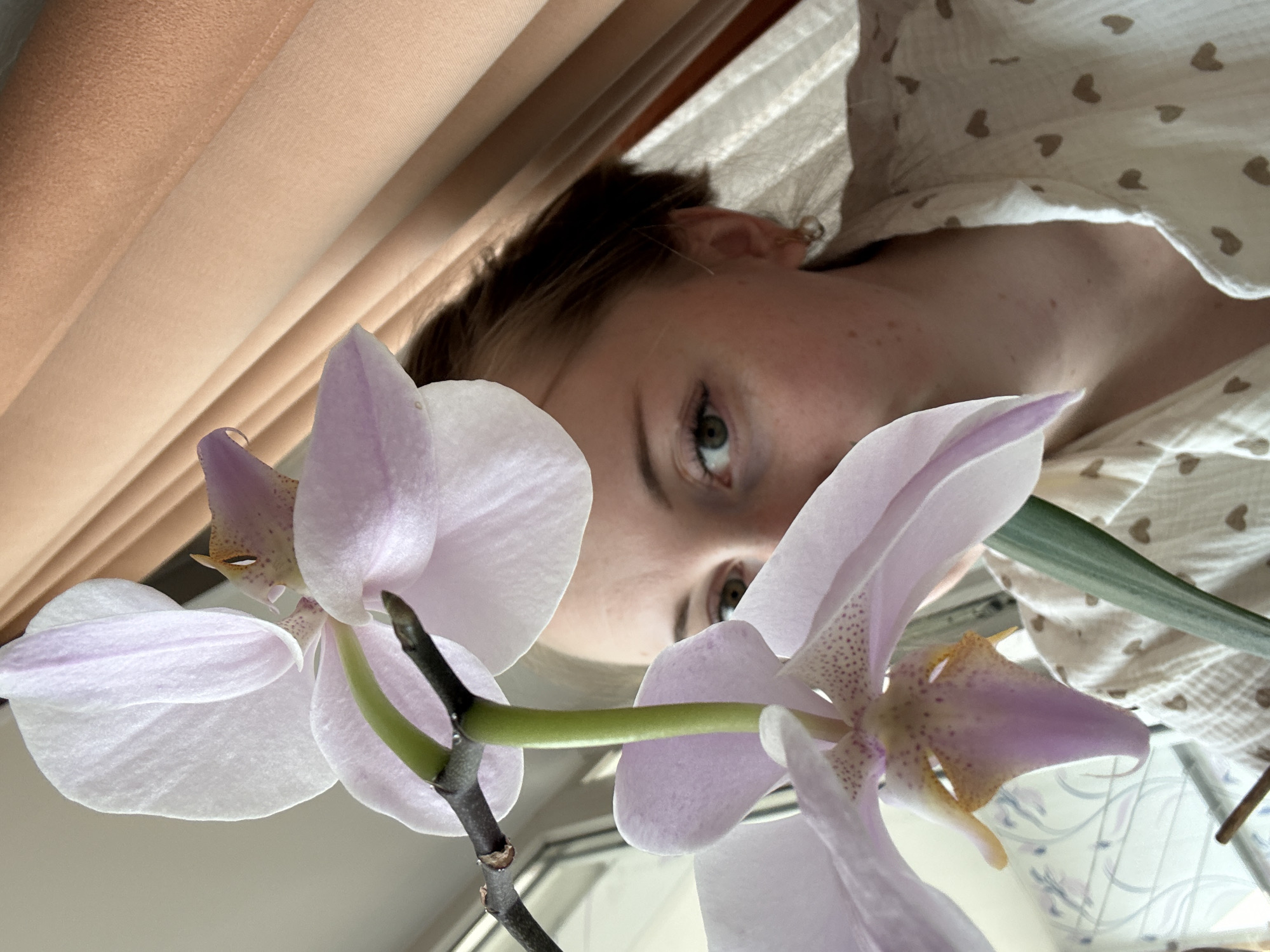 A close-up, tilted photograph shows a person partially obscured by two light purple orchid blossoms. The person, whose eyes are in focus, has fair skin with freckles. They are wearing a white top with small brown heart prints. The orchid blossoms are in the foreground, with a slender green stem visible. In the background, a tan curtain hangs diagonally, and a window with a patterned blind or curtain is partially visible. The location is Unknown, Unknown.