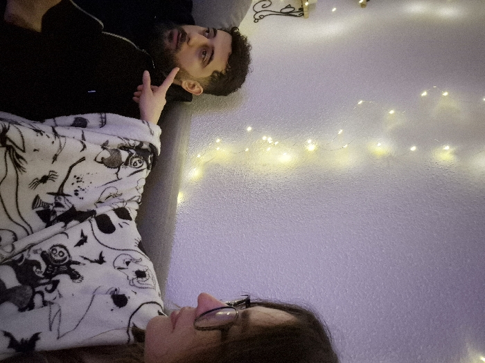 An indoor scene in Strasbourg, France, depicts two individuals seated on a grey upholstered couch. On the left, a male subject with dark curly hair and a beard, wearing a black hooded jacket, is shown in profile looking towards the left. To his right, a female subject, partially visible, wears glasses and is covered by a white blanket featuring black Halloween-themed patterns such as ghosts, bats, and skulls. Her right index finger is extended, pointing towards the male's left cheek/jawline area. The background consists of a light-colored, textured wall adorned with vertically strung warm yellow fairy lights. A dark, ornate wall bracket is partially visible on the far left.