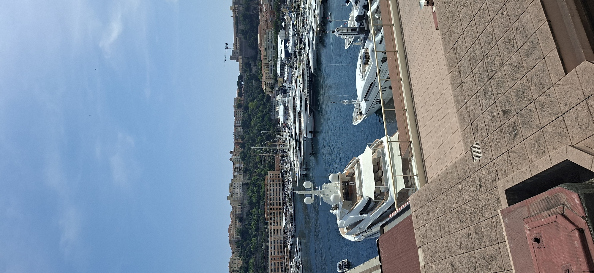 The image depicts a panoramic view of the Port Hercules in Monaco on a clear, sunny day. The foreground features a tiled promenade. In the middle ground, a large white yacht is docked prominently, with a smaller boat to its left. The harbor is filled with numerous other yachts and boats of various sizes, all moored in the blue water. In the background, the cityscape of Monaco rises, with buildings constructed on a hillside covered in lush green trees. At the very top of the hill, a fortress-like structure with a crane nearby is visible, likely the Prince's Palace of Monaco. The sky is a bright, clear blue with a few wispy clouds and two distant birds.