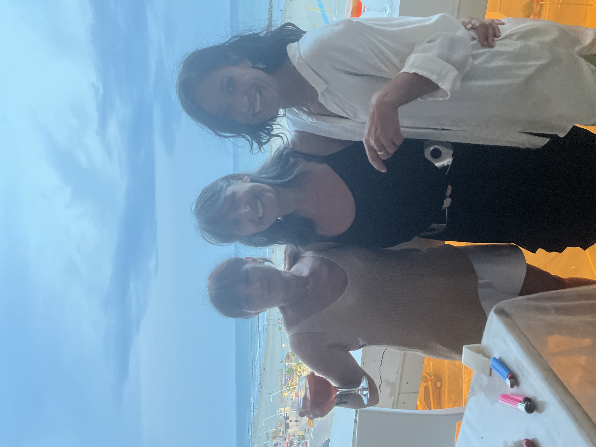 Three women stand on a balcony overlooking a beach and the Mediterranean Sea. The woman on the left, with short brown hair, holds a glass of pink cocktail in her right hand and wears a tan sleeveless top and white shorts. The woman in the middle, with shoulder-length graying brown hair, wears a black sleeveless dress with a silver belt and smiles. The woman on the right, with dark wavy hair, wears a loose white shirt and smiles as she places her left hand on the middle woman's arm. A table in the foreground is covered with a white cloth, and two lighters, one pink and one blue, sit on it. The scene appears to be in Palavas-les-Flots, France, as suggested by the architecture and beach layout resembling the area. The sky is overcast with a pale blue hue.