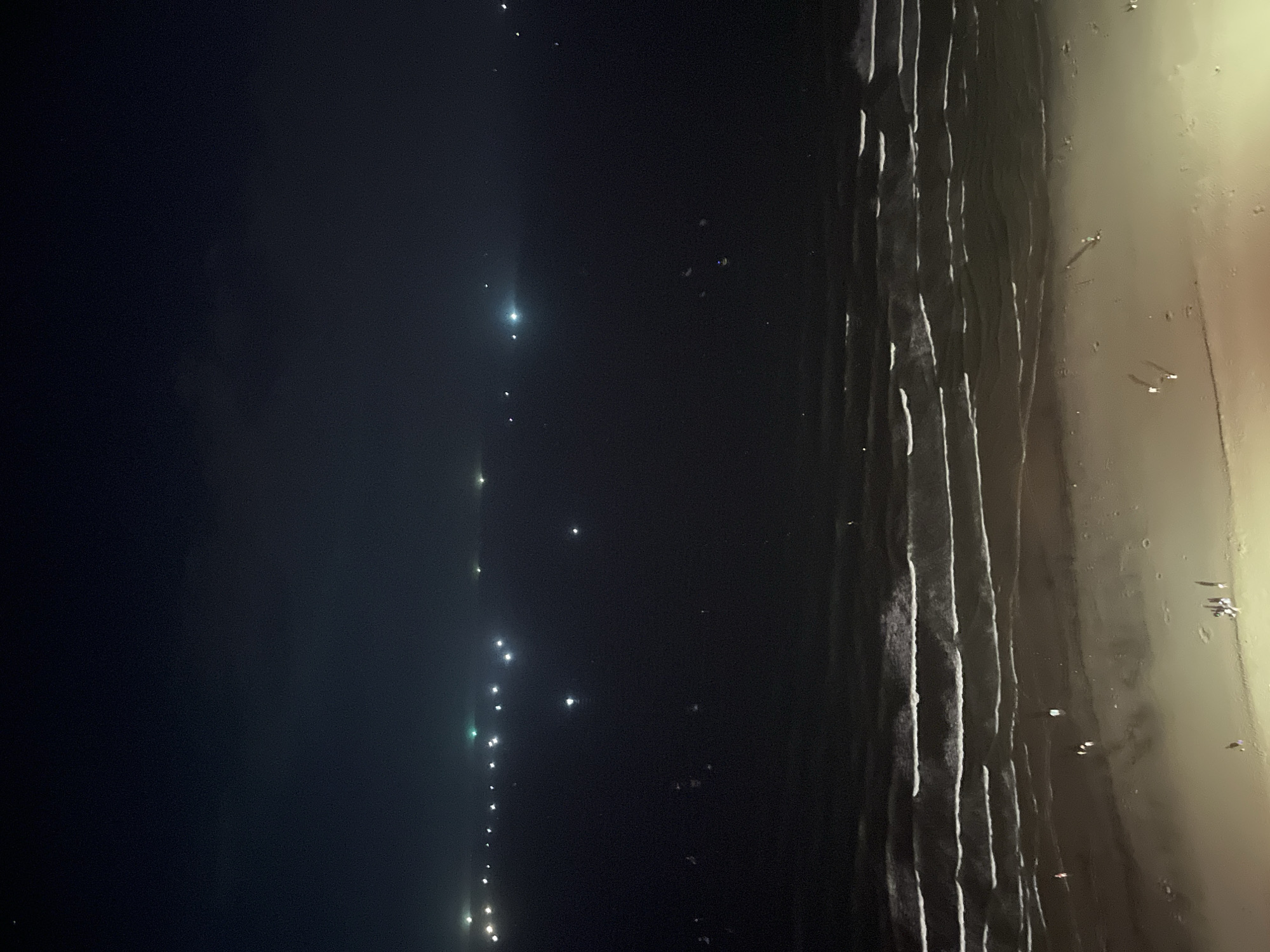 This is a high-angle, nighttime photograph of a beach in Da Nang, Vietnam. The scene is dimly lit by artificial lights. The ocean is on the left and center, with gentle waves breaking on the shore. The beach is on the right and bottom, with some scattered figures, likely people, walking or standing. In the distance, across the water, are numerous small, bright lights, possibly from boats or a distant coastline. The sky is dark and appears to have a slight haze.