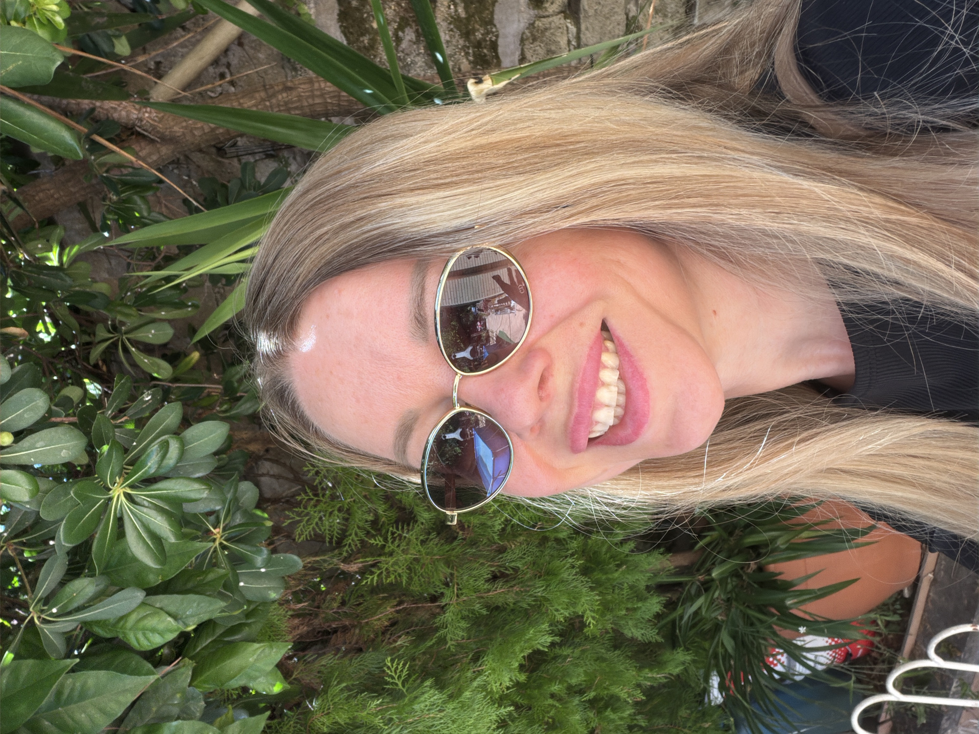 A medium shot shows a woman with blonde hair and sunglasses smiling outdoors. She is wearing a black top. The background consists of lush green foliage and a stone wall. The sunglasses reflect a scene that appears to be a courtyard or patio with furniture. The lighting suggests it is daytime.