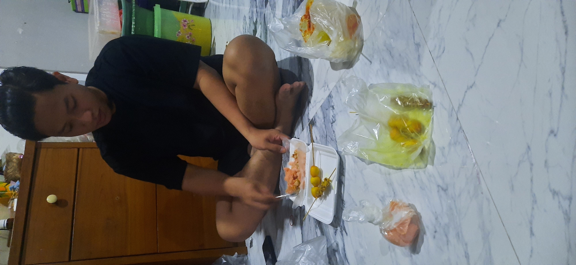 A young adult male is sitting cross-legged on a marble-patterned floor, wearing a black t-shirt. He is looking down at food that is spread out on the floor in front of him. The food appears to be in various plastic bags and a white styrofoam container. There are at least three skewers with food items, as well as some pinkish food in a plastic bag. In the background, there is a wooden dresser with a single knob and some containers on top. The scene suggests a casual meal or snack at a residence.