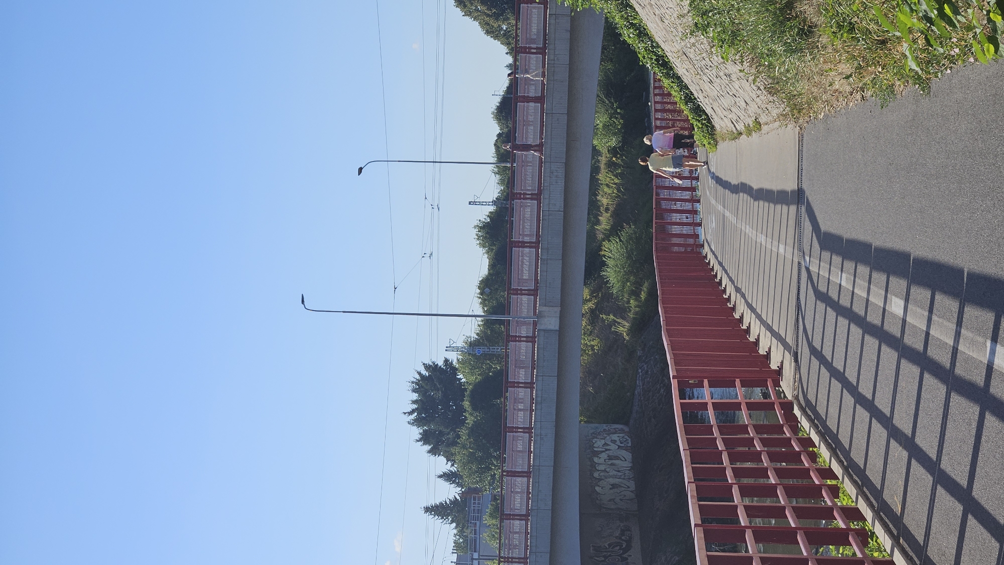 Two women are walking on a paved path next to a body of water, with a red metal fence separating them from the water. The path has a white line marking a bike lane. In the background, there is a bridge with people walking on it. Trees and vegetation are visible on the sides of the path and under the bridge. The sky is clear and blue. This scene is located in Prešov, Slovakia.