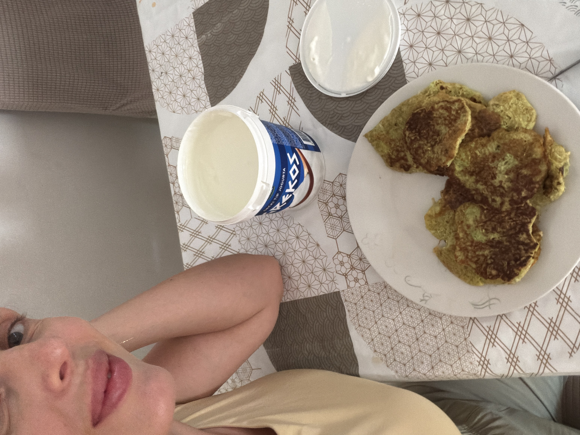 The image shows a close-up view of a person's face and arm, along with a plate of food and a container of yogurt. The person is lying down, with their head propped on their arm and their tongue sticking out slightly. They appear to be in a relaxed or playful mood. The food on the plate consists of several small, circular, greenish-brown pancakes, possibly zucchini or spinach fritters. Next to the plate is an open container of white yogurt, with its lid placed beside it. The items are arranged on a table covered with a patterned tablecloth. The tablecloth features a geometric design with brown and white elements. In the background, a brown upholstered piece of furniture, possibly a sofa, is visible. The lighting suggests it could be daytime, with natural light illuminating the scene. The overall setting appears to be a casual indoor environment, likely a home or a rented accommodation. The visible text on the yogurt container reads "KEKOS YOGURTA". The context provided indicates the location is Budva, Montenegro.