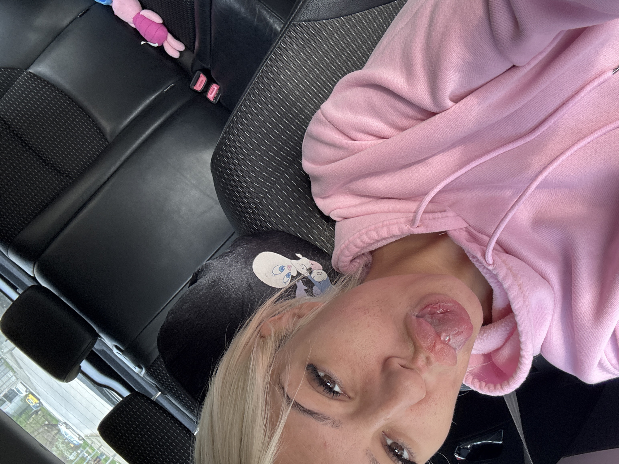 A close-up, selfie-style image appears to have been taken from inside a car. The camera is angled downwards towards a woman with blonde hair wearing a pink hoodie. She is sticking her tongue out. The car's interior is visible, including black leather seats. In the background, a glimpse of the outside world is visible through the window. The location, however, remains unknown.