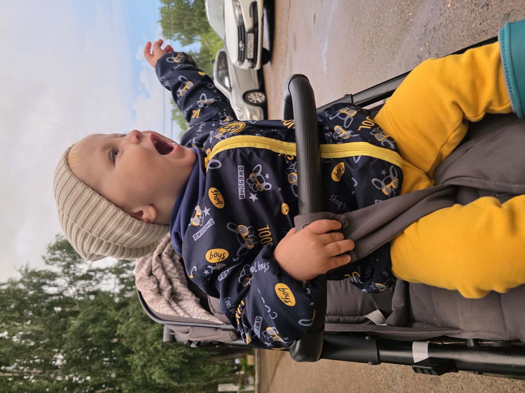 A light-skinned toddler wearing a beige knit cap, a dark blue jacket with a yellow zipper and bee-themed pattern, and bright yellow pants sits in a gray stroller. The toddler is facing to the right, with their mouth open in a yawn or shout, and their right arm is extended outwards. In the background, there are trees and two cars parked on a wet asphalt surface. The sky is overcast with some blue sky visible. The scene appears to be outdoors, possibly in a residential area or park in the city of Chaikovsky, Russia.