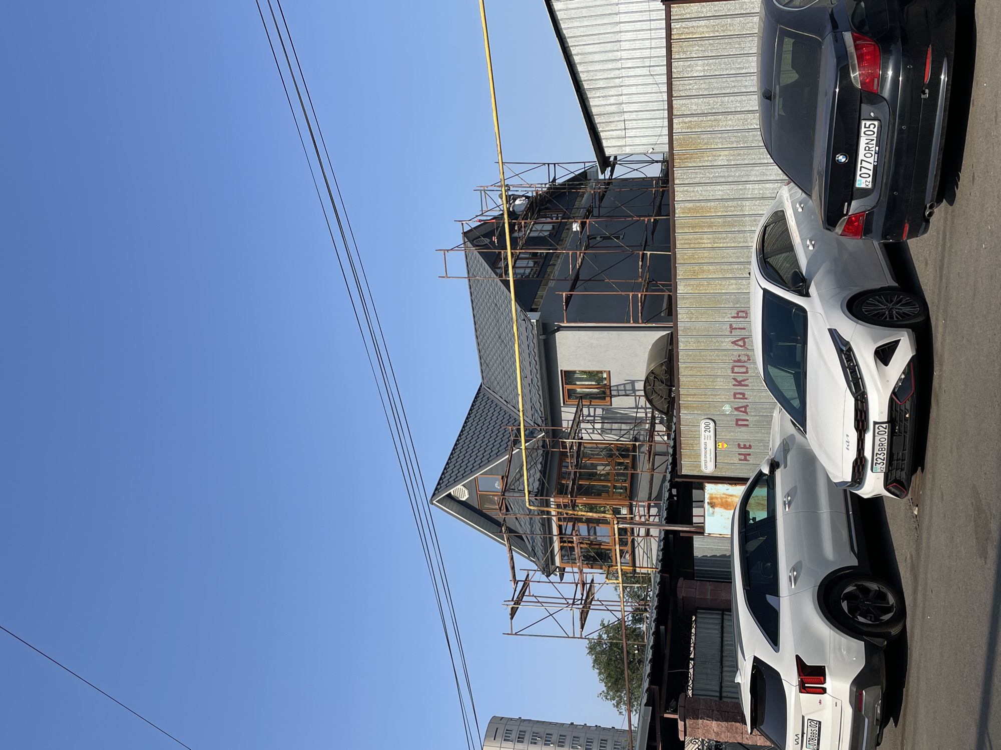 In Almaty, Kazakhstan, on a clear day, a residential house under construction is visible, surrounded by scaffolding. A yellow gas pipeline runs horizontally across the frame in front of the house. To the left of the house, a tall, modern apartment building is partially visible. In the foreground, three cars are parked on a paved surface: a white Kia SUV, a white Kia sedan, and a black BMW sedan. A metal fence with corrugated siding runs behind the cars and in front of the house. A sign on the fence, partially obscured by the cars, reads "НЕ ПАРКОВАТЬ" (Do Not Park) in Cyrillic letters. The overall scene depicts an urban residential area with ongoing construction and parked vehicles.