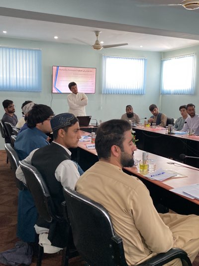 The image depicts a group of men in a room in Shurandam, Afghanistan. The men are seated around a long table, and one man is standing at the front of the room giving a presentation. A screen behind him displays text. The room appears to be a conference or meeting room, with blinds on the windows, a ceiling fan, and a brown carpet. There are glasses of water and papers on the table. The men are dressed in traditional clothing.