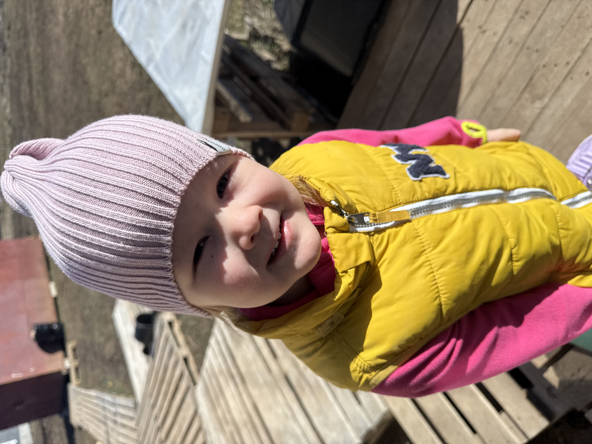 A young child, likely a toddler, is the central subject of the image. The child is wearing a pink knit hat and a yellow puffer vest over a pink long-sleeved shirt. The vest has a zipper and a patch on the chest. The child is smiling slightly, showing their teeth.
The background consists of a wooden structure, possibly a deck or platform, with gaps between the planks. In the distance, there are hints of other structures and vegetation, including what appears to be a white tarp.
The photo was taken outdoors in Gornyi (Dvurechenskoe S/P), Russia.
