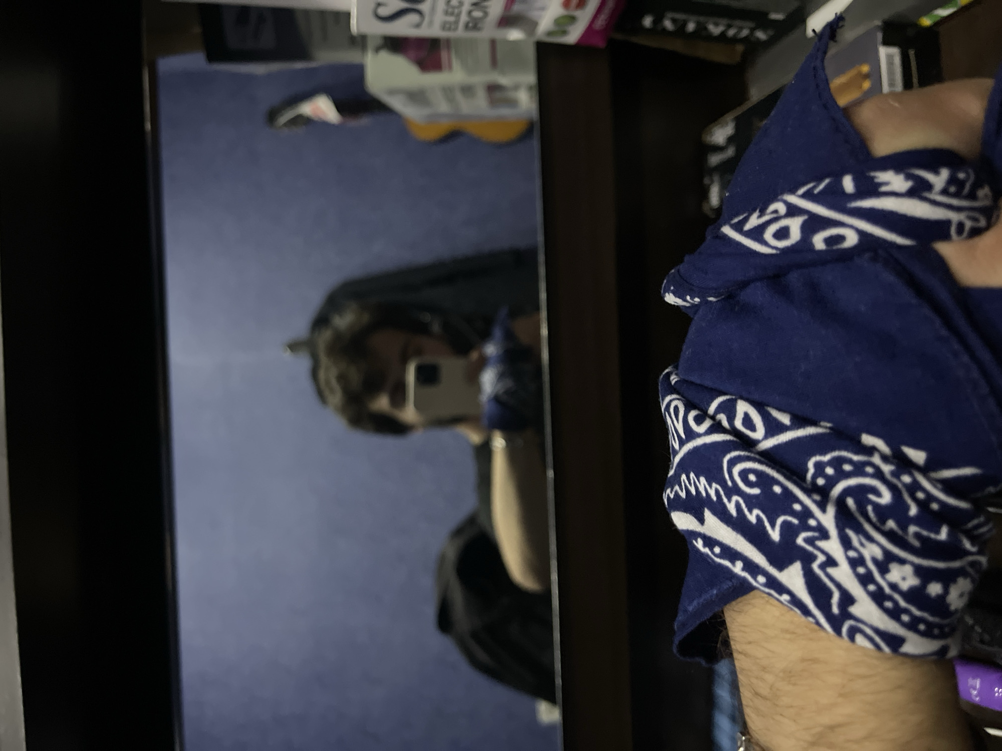 The image is a close-up, low-angle shot of a person's arm and wrist wearing a blue bandana with a white paisley pattern. The arm is hairy and positioned in the foreground. In the background, reflected in a mirror, a person is taking a selfie with a smartphone. The reflected person has dark, curly hair and is wearing a dark shirt. The background in the mirror shows a plain blue wall, a guitar, and shelves with various objects. The overall setting appears to be an indoor space, possibly a room or a studio. There is no discernible information suggesting the location is the city of Tbilisi, Georgia.