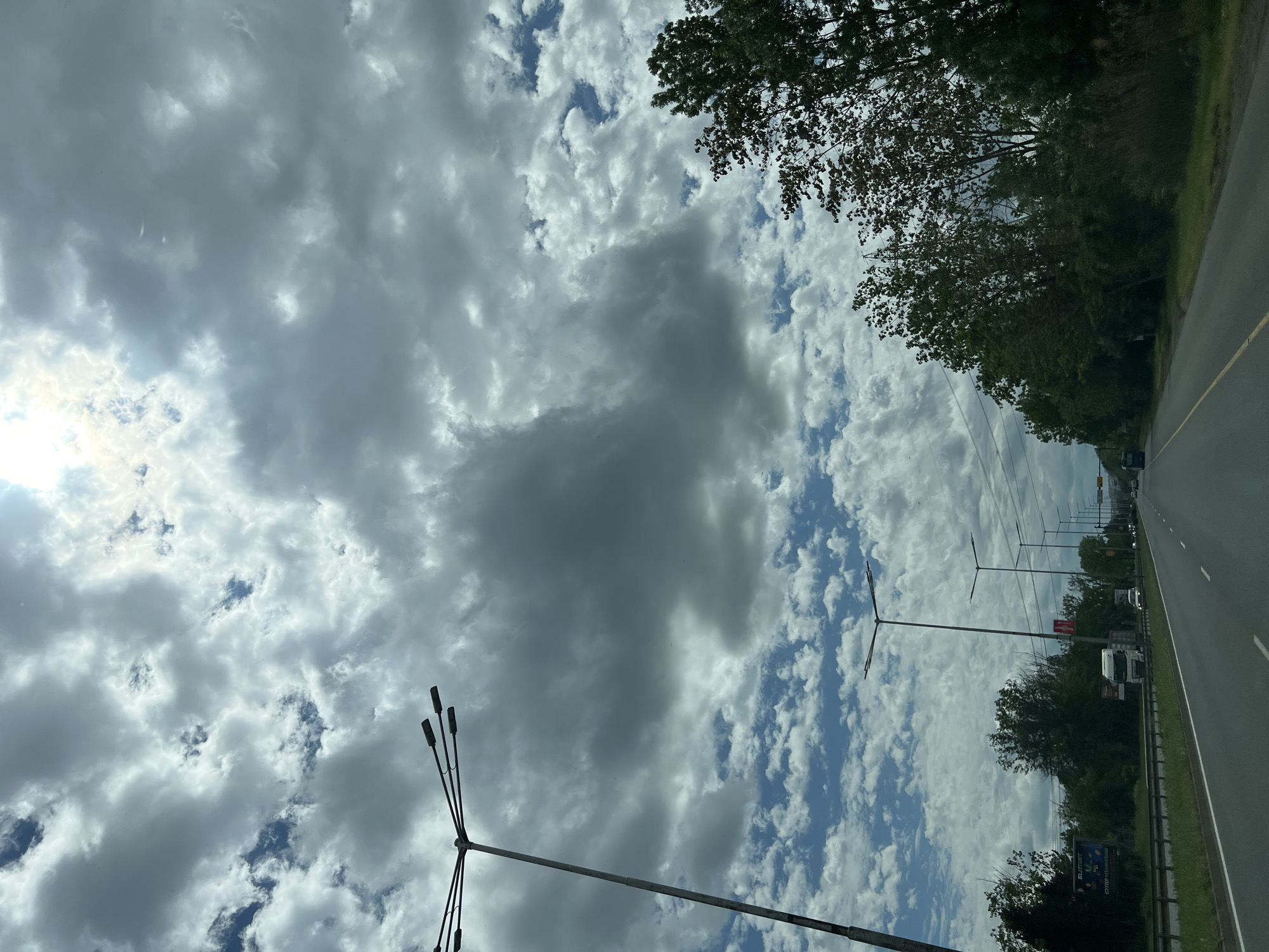 The image depicts a view from inside a vehicle, likely a car, traveling on a road in what appears to be a city or suburban area. The sky is mostly cloudy with patches of blue visible. There are trees lining the side of the road. Street lights are visible on the side of the road. There are vehicles further down the road. The image does not allow the determination of the location within Burgas, Bulgaria. There are no people visible.