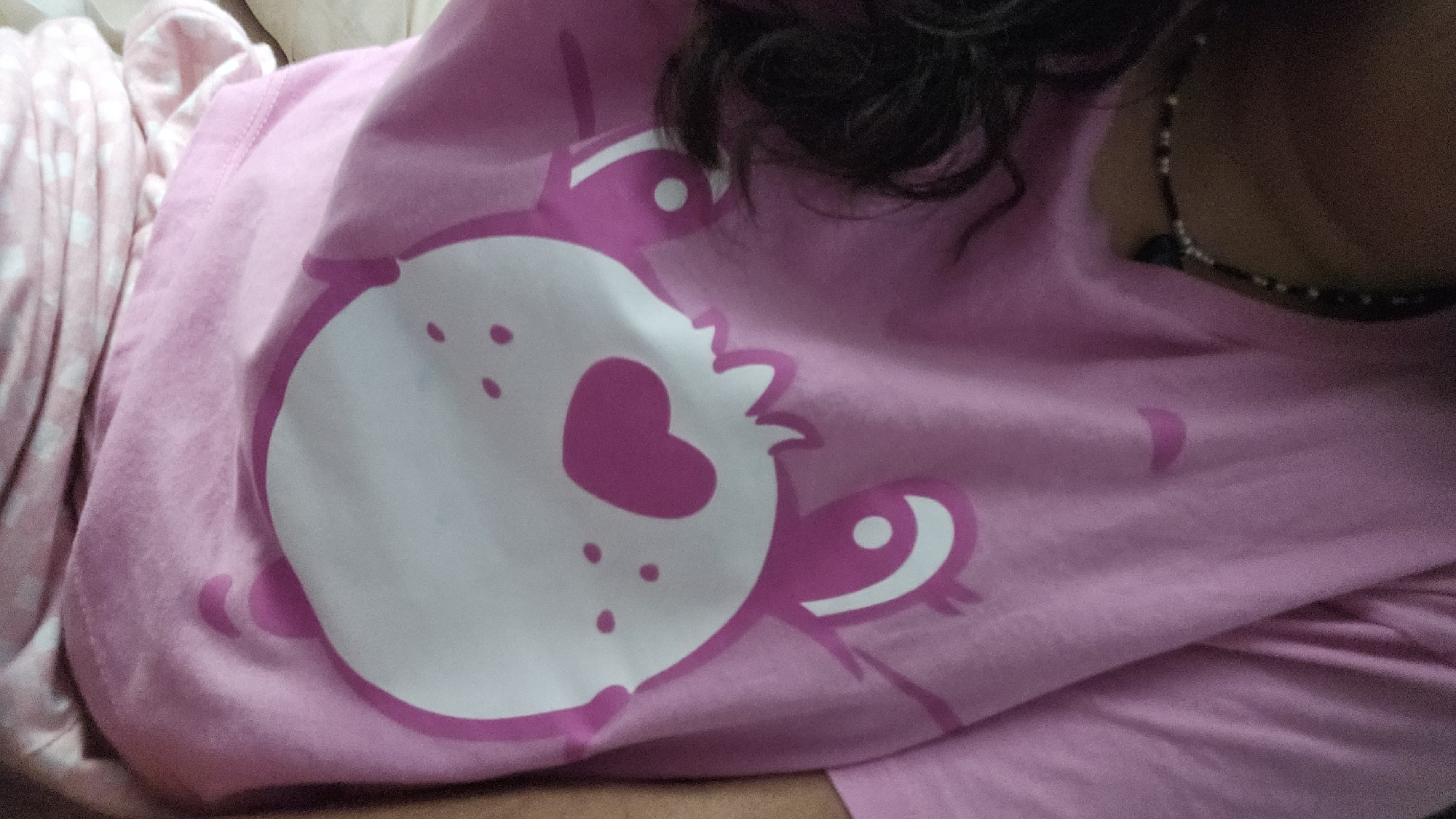 A close-up shot captures a person wearing a light pink t-shirt adorned with a white and darker pink stylized cartoon crab design, featuring a heart shape on the crab's body. The subject is also wearing a beaded necklace. Part of the subject's curly dark hair is visible. To the left, there are partially visible patterned pajamas. The exact location of the photo cannot be determined from the visual context.