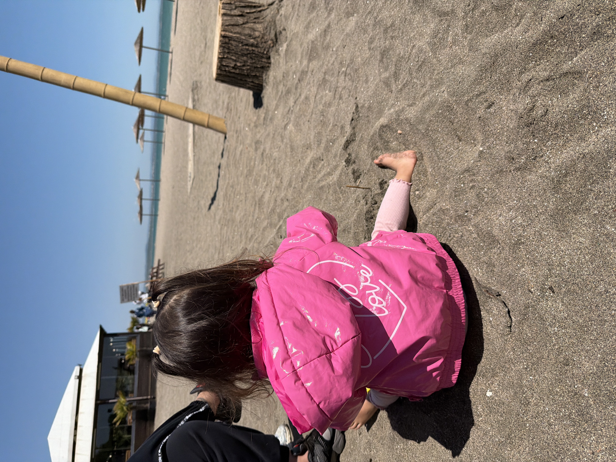 A girl is playing on a sandy beach. She is wearing a bright pink jacket. The sand is dark. In the distance, there are beach umbrellas and a bamboo structure. In the background, there is a building and people. The scene is well-lit with clear sky.
