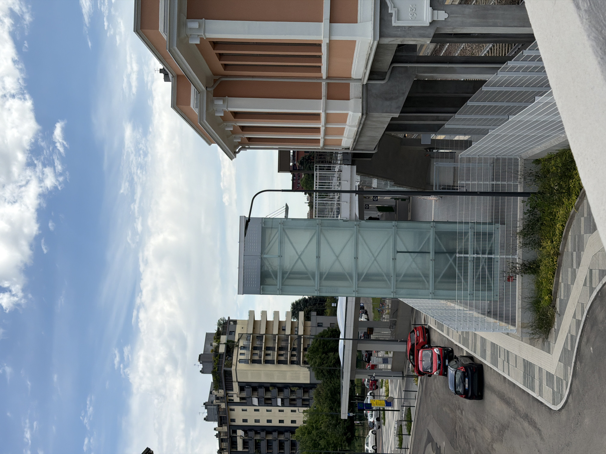 The image depicts an urban scene, likely in Milan, Italy. The view is from an elevated position, looking towards a street.
Architecture: A building with peach-colored exterior and white trim is visible at the top of the frame. It appears to be an older style building. Modern architecture is also present including a glass enclosed structure and apartment blocks.
Street Scene: The street features a curb and road markings. Several parked cars are visible in red and black. There's some signage and street furniture (lampposts).
Vegetation: Green plants and grass are incorporated into the urban landscape.
Sky: The sky is visible in the background, with a mix of blue sky and scattered clouds.